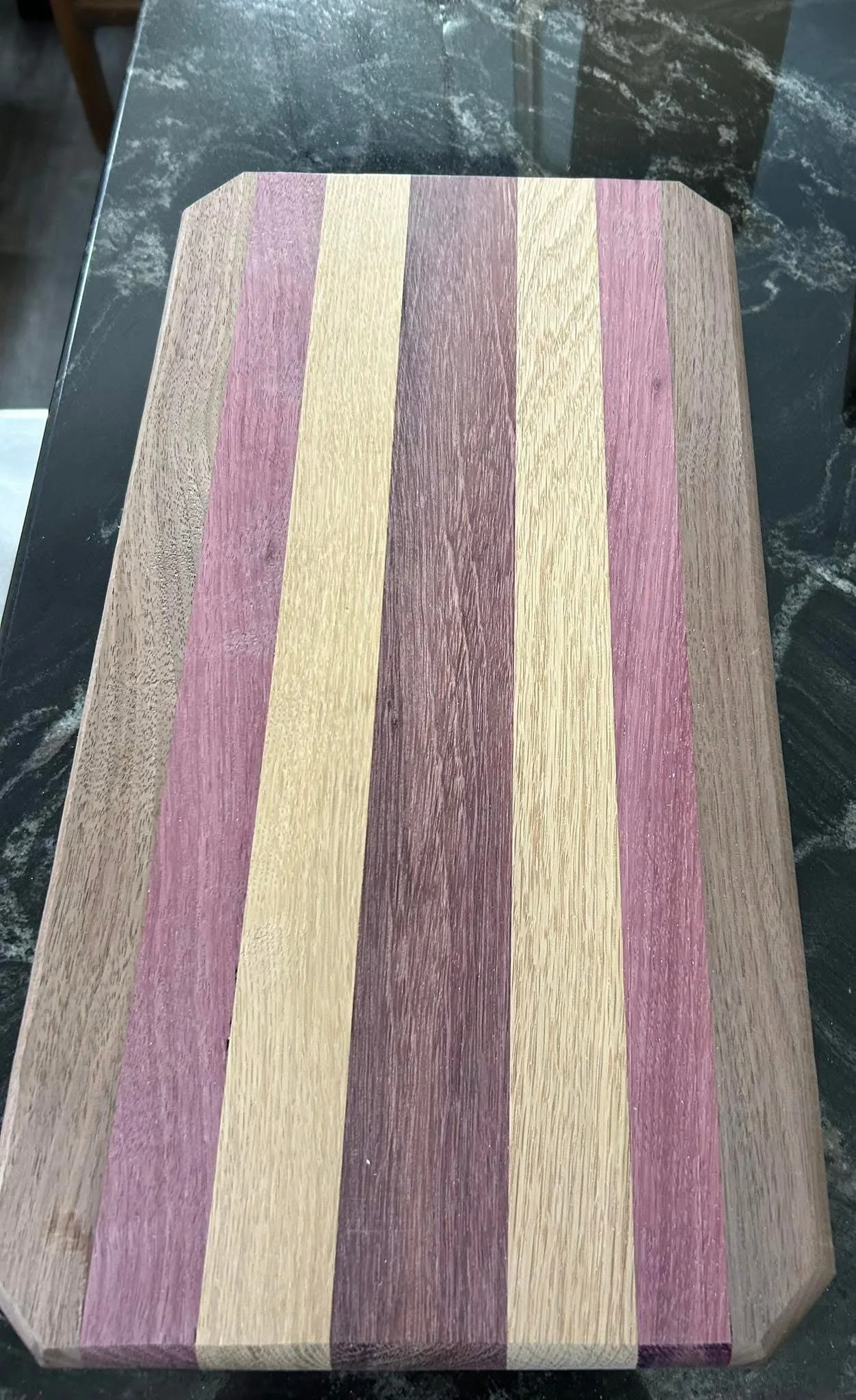 Wooden cutting board with multicolored strips of purple, beige, and brown, on a black marble countertop.