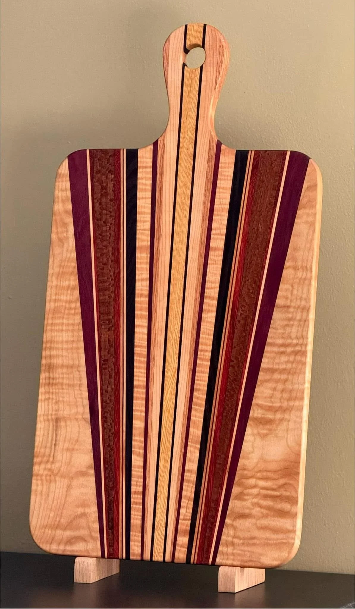 Wooden cutting board with multicolored stripes used for serving or chopping, standing upright on a black surface against a beige wall.