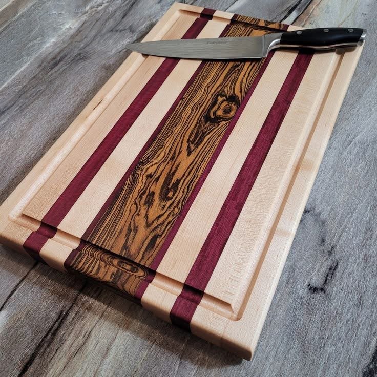 Wooden cutting board with red and light wood stripes, and a chef's knife placed on top, on a wooden surface.
