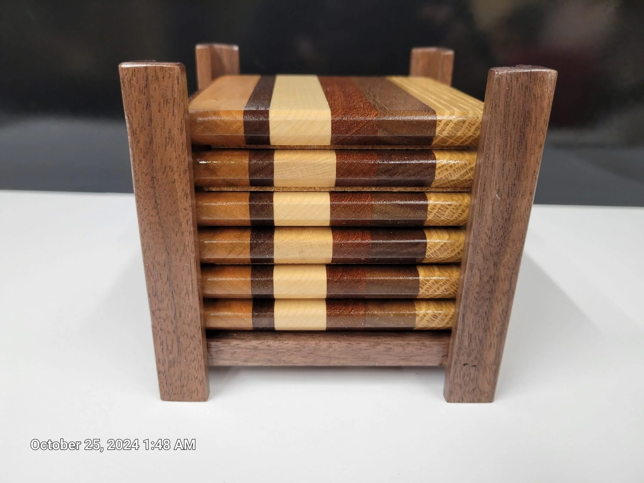 Stack of four wooden coasters with square patterns in light, dark, and medium wood tones, held within a wooden holder with four sides, placed on a white surface.