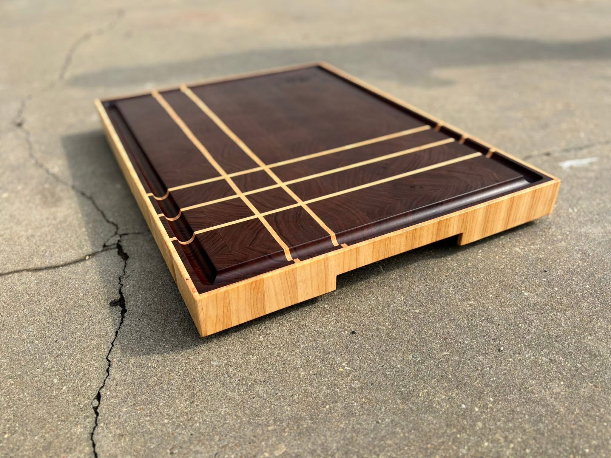 A wooden game board with a sleek, dark brown surface and light wood grid lines, lying on a concrete pavement.