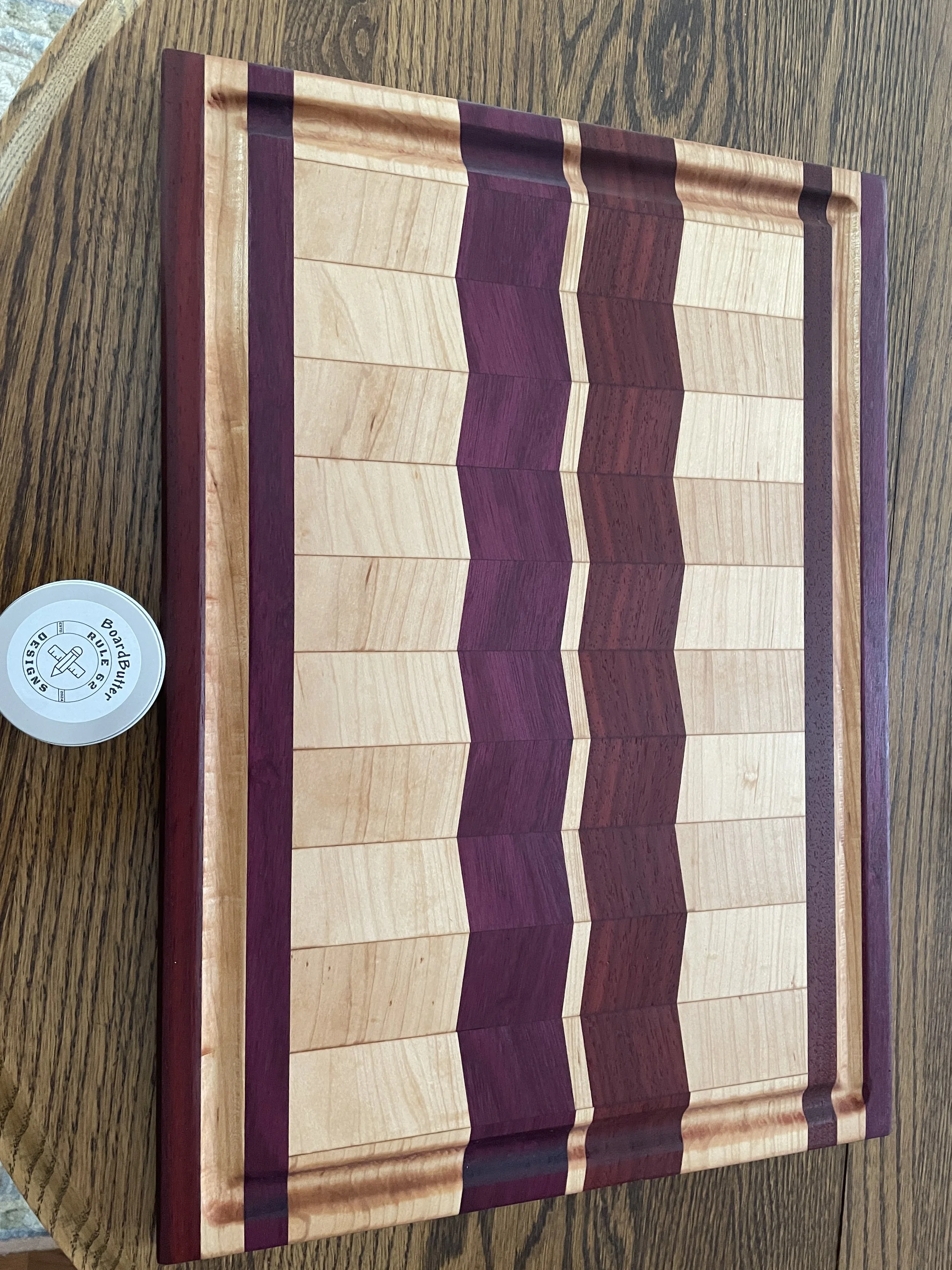 A wooden chessboard with alternating light and dark purple wood pieces arranged on a wooden table. A small container labeled 'Board Butter' sit next to the board.
