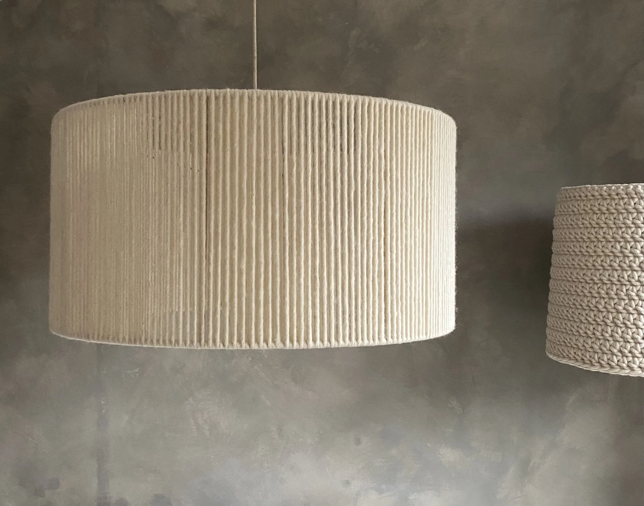 Two hanging fabric lampshades, one large beige with vertical ribbed texture, and one smaller beige with a woven texture, against a gray wall.