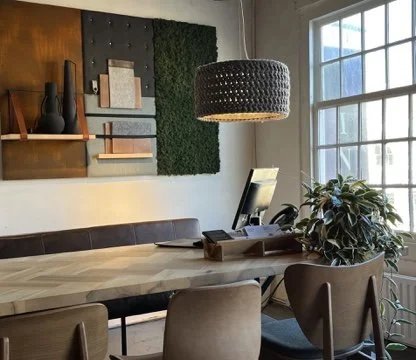 A modern office space with a large table, a computer monitor, a potted plant, and a stylish hanging light fixture near a window.