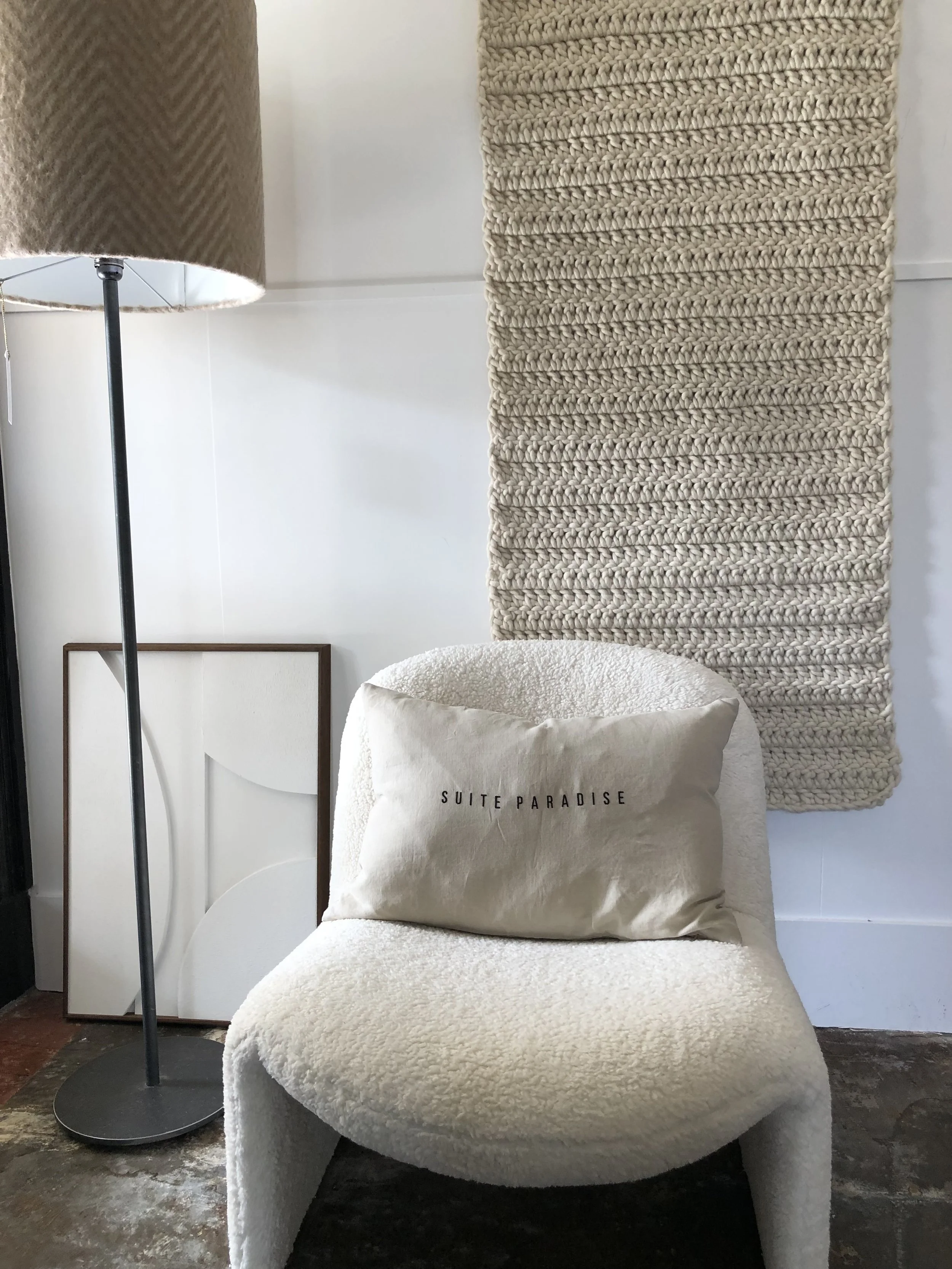 A cozy corner with a floor lamp, a framed artwork leaning against the wall, a textured beige wall hanging, and a plush white chair with a beige pillow that reads "SUITE PARADISE" in black letters.
