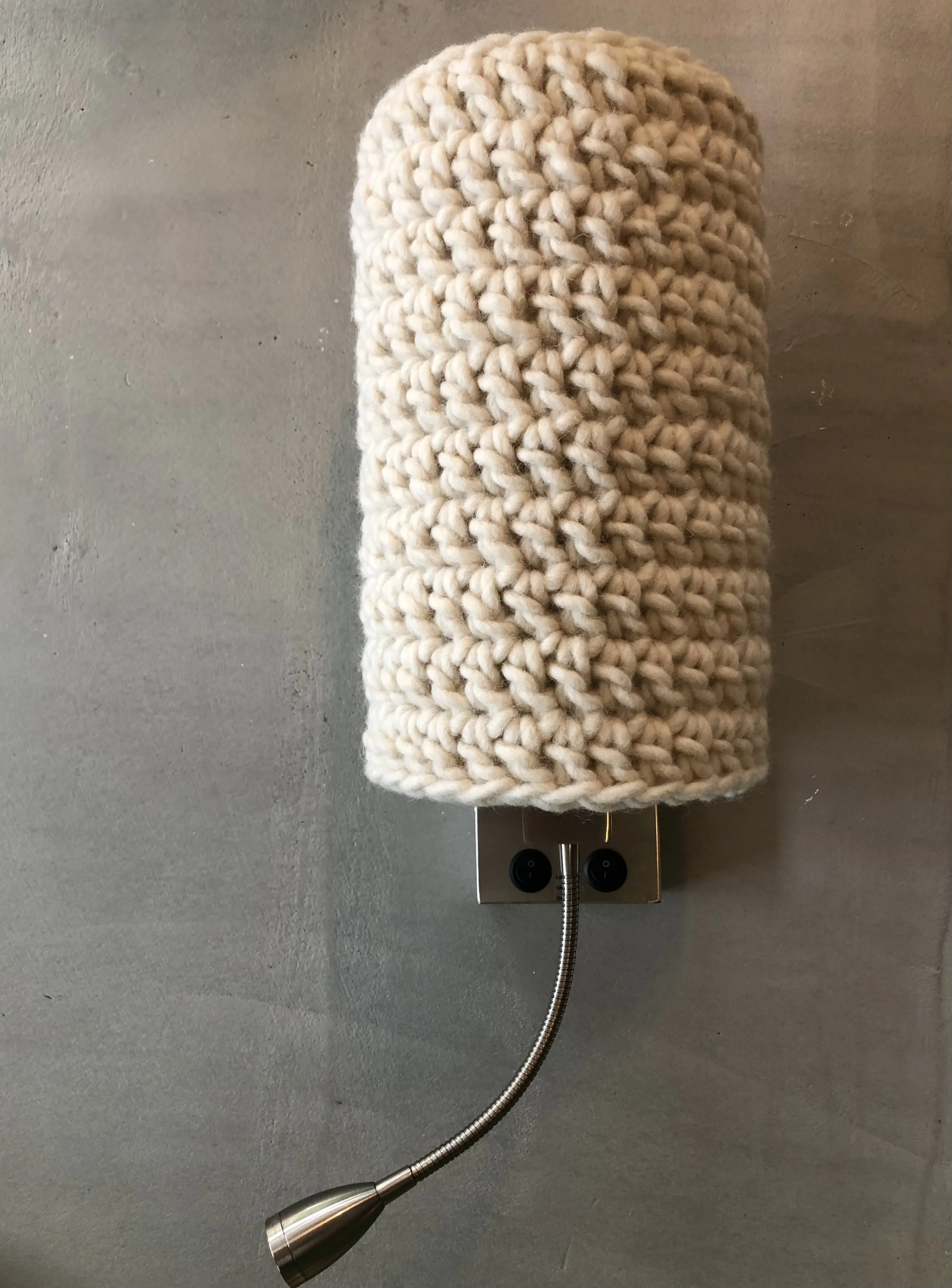 A wall-mounted lamp with a flexible neck, covered by a thick, cream-colored, knitted cover, situated on a light gray wall.
