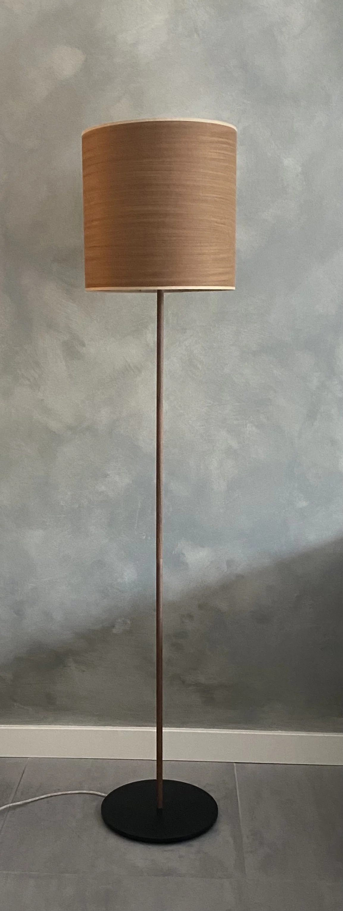 A floor lamp with a black round base, a thin wooden pole, and a large cylindrical beige lampshade, placed against a gray textured wall.