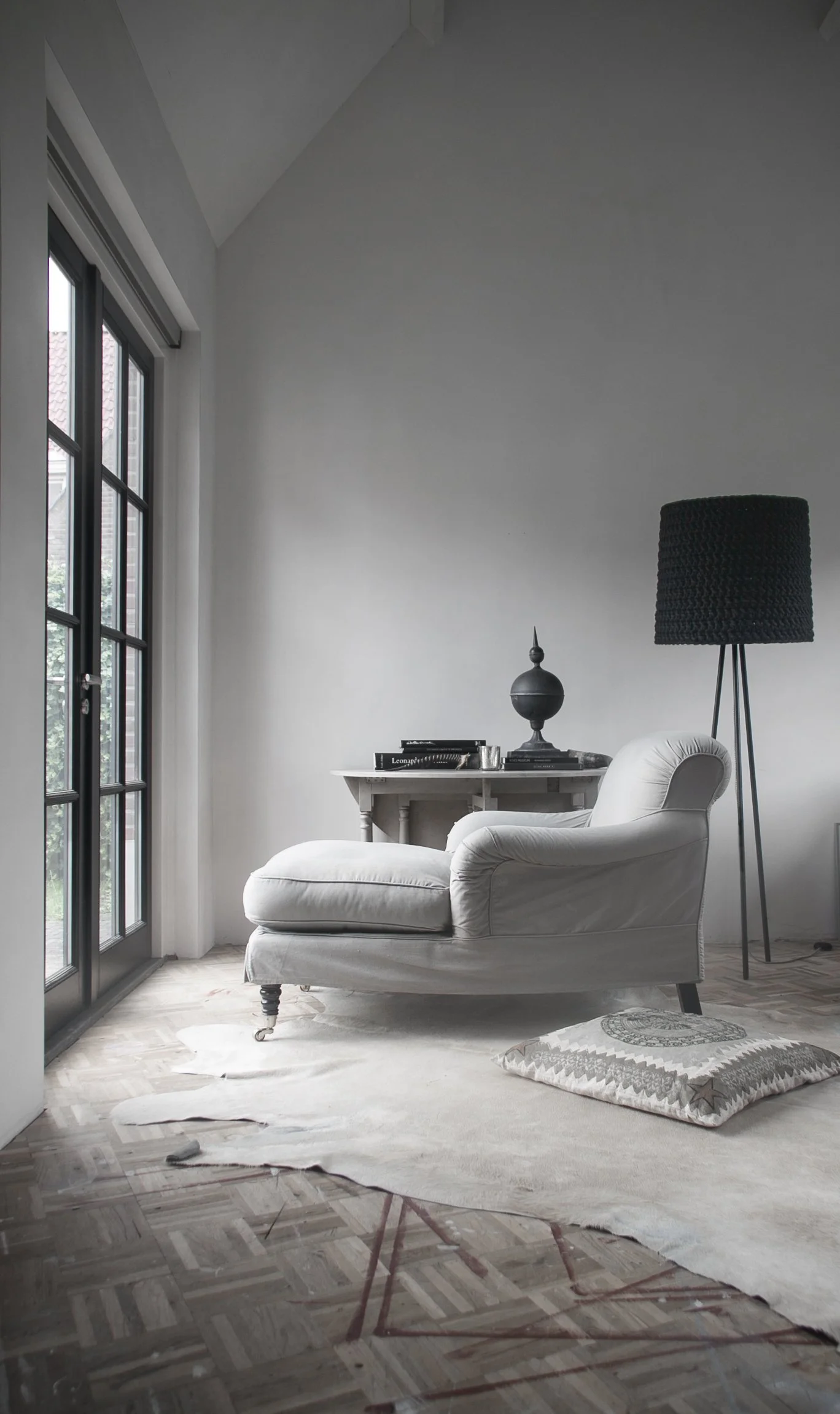 A white chaise lounge chair in a bright room next to a large black floor lamp and a small white side table with decorative objects, window with black frame showing an outdoor view, and a patterned rug on parquet flooring.