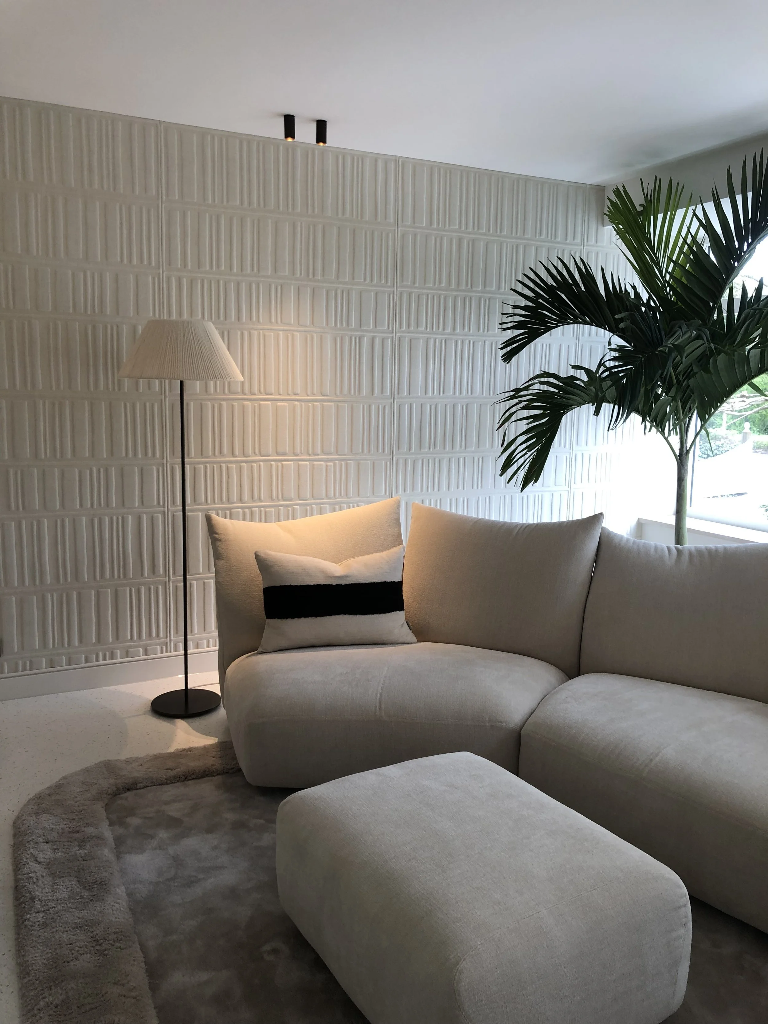 Living room with a beige sectional sofa, a matching ottoman, a black and beige pillow, a tall floor lamp, a large potted palm tree, cream-colored textured wall, carpet, and window with natural light.