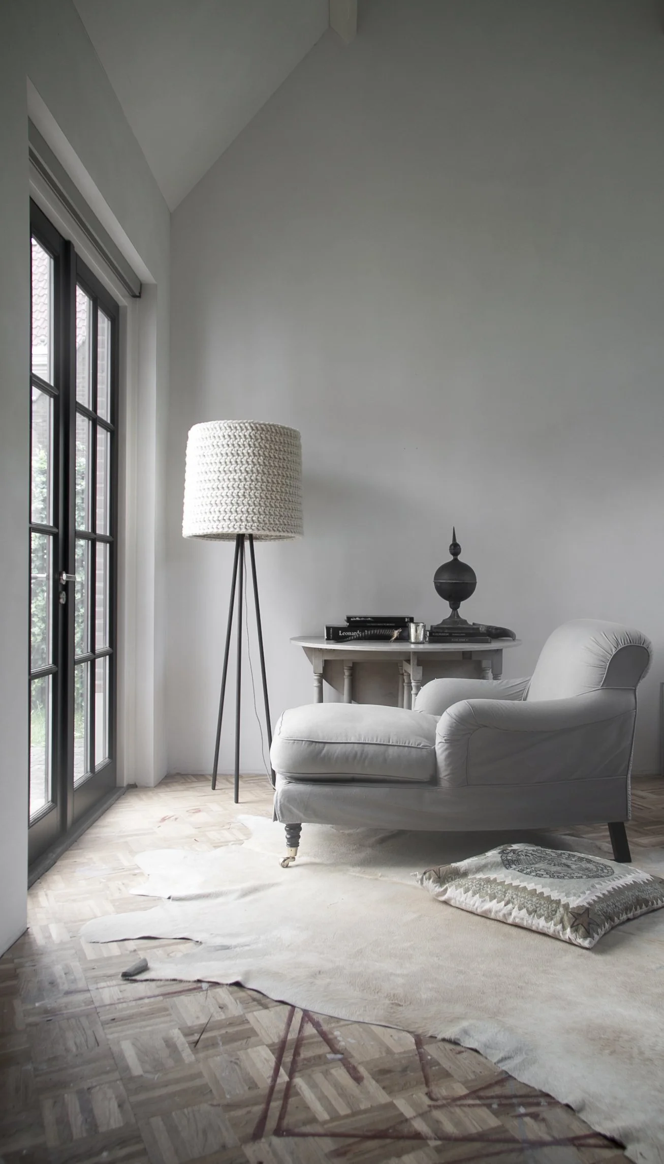 A cozy living room corner with a large floor lamp, a white armchair with a matching ottoman, a small side table with books, a black decorative object, and a floor cushion on a white rug near glass doors leading outside.