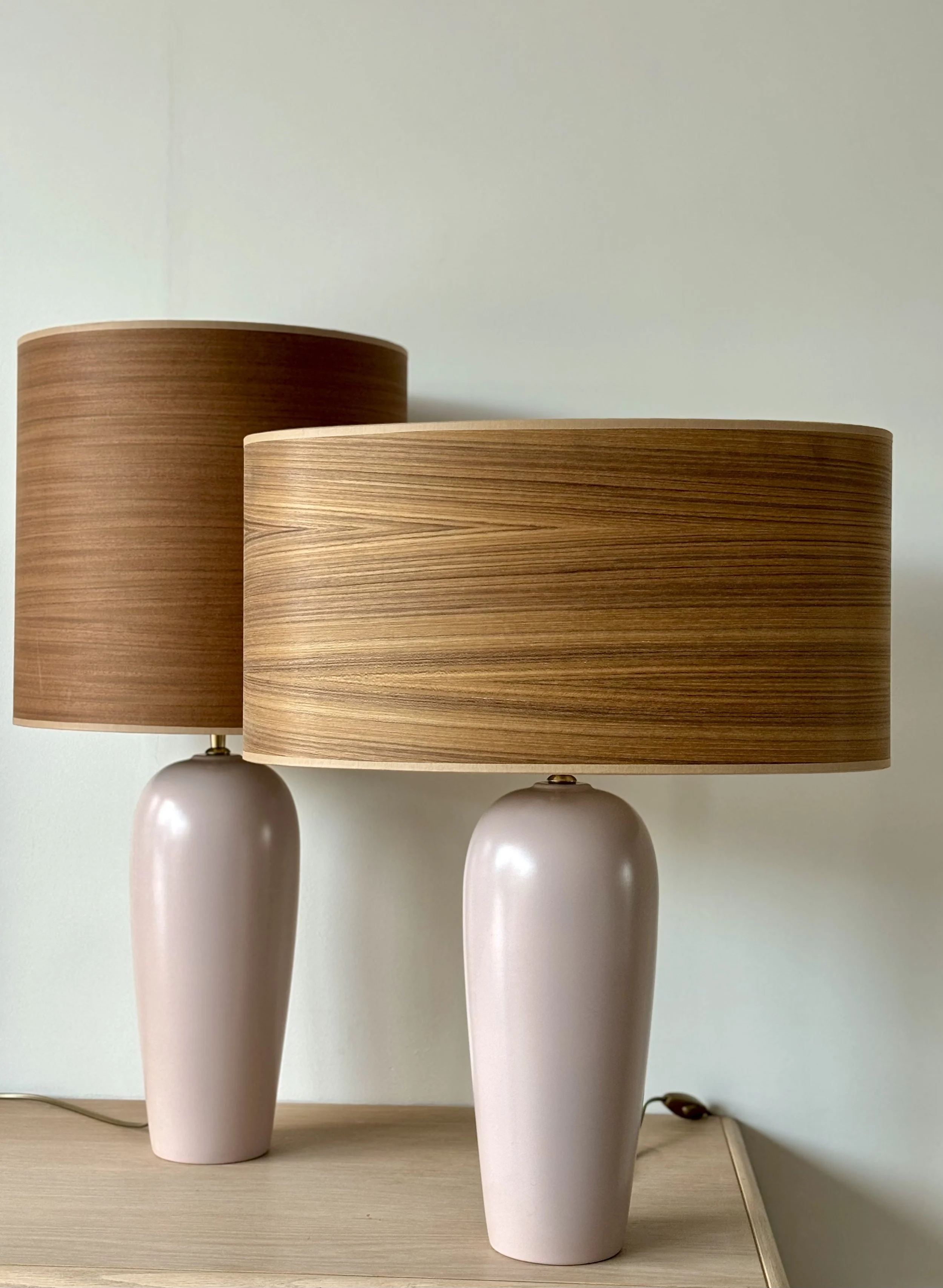 Two table lamps with oval pink ceramic bases and large wooden cylindrical shades on a light wood surface against a plain wall.