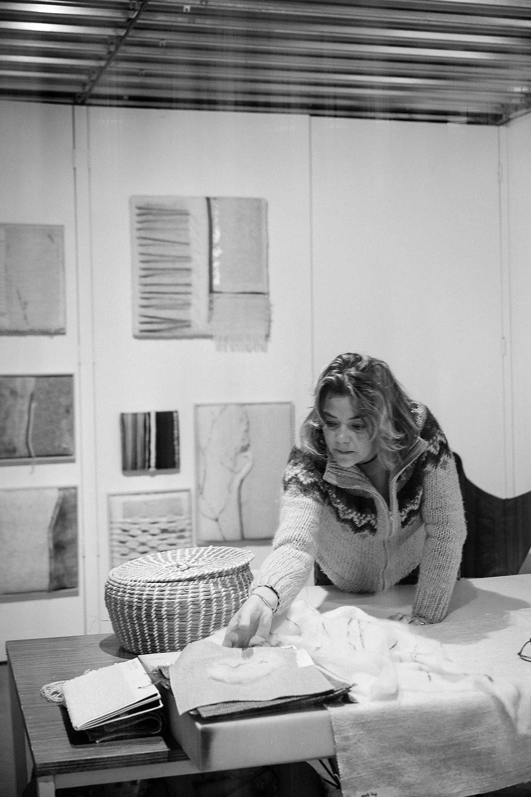 A woman in a knit sweater is leaning over a table, organizing or sorting fabrics and papers, with artwork and textiles displayed on the wall behind her.