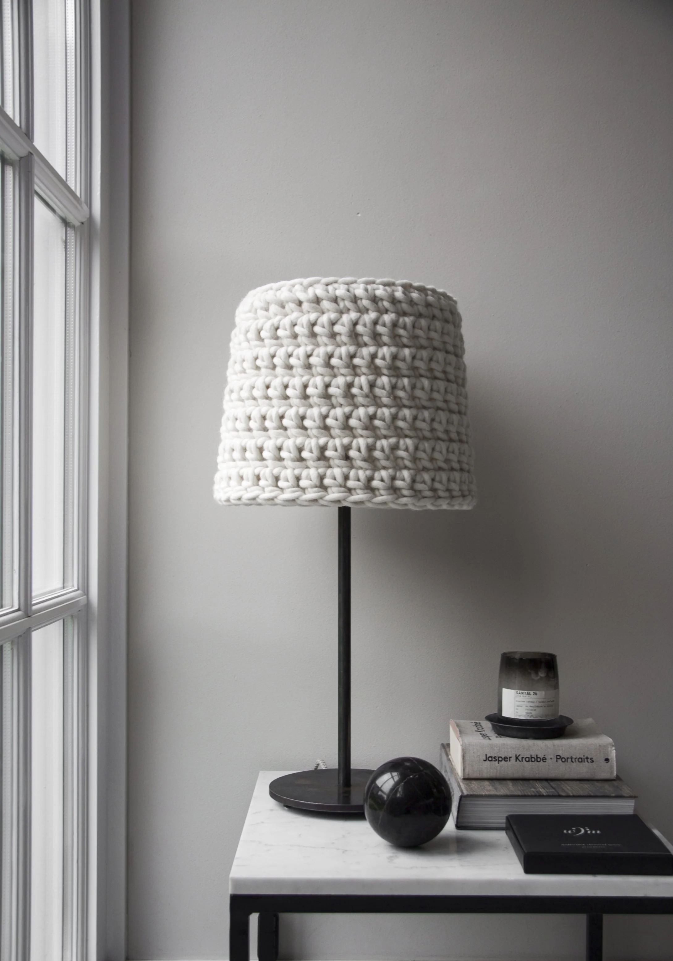 A white woven table lamp on a white marble side table, next to books, a black vase, and a black candle holder, with a window on the left.