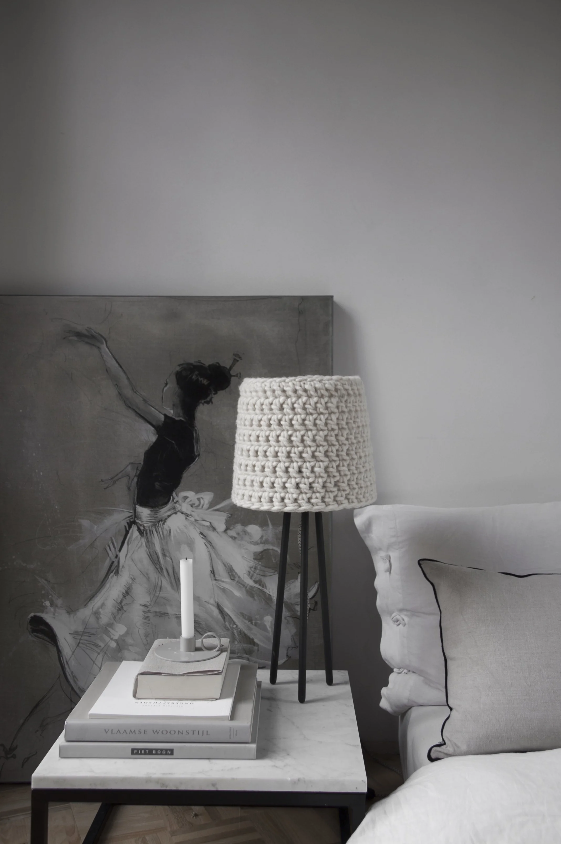 A modern bedroom corner with a white marble side table, a stack of books, a small candle, and a table lamp with a thick, textured, neutral-colored shade. Behind the table, there is a large painting of a woman in a dance pose, and to the right, part o