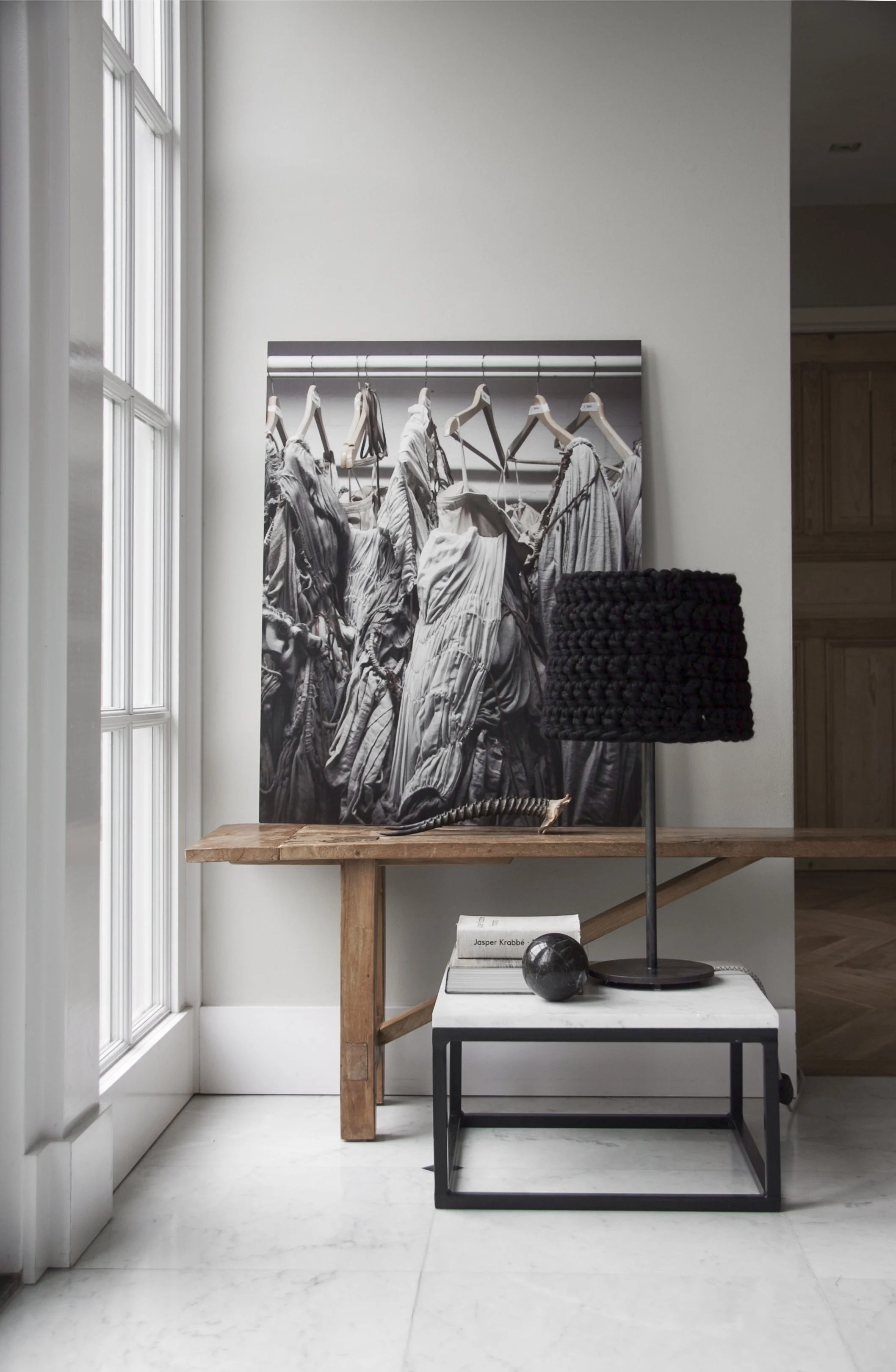Interior corner with a window on the left, a black-and-white artwork of hanging garments on the wall, a wooden table, a small black-and-white side table with a book and a decorative black sphere, and a black textured lamp.