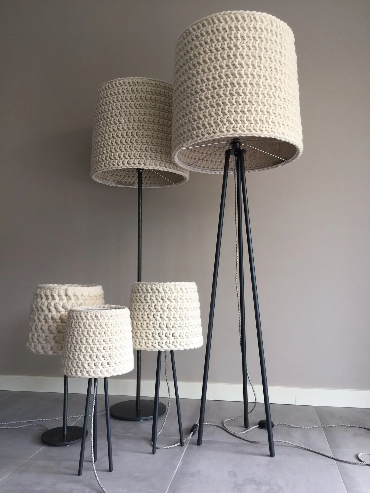 Set of five floor lamps with knitted cream-colored lampshades and black tripod bases, arranged against a plain gray wall.
