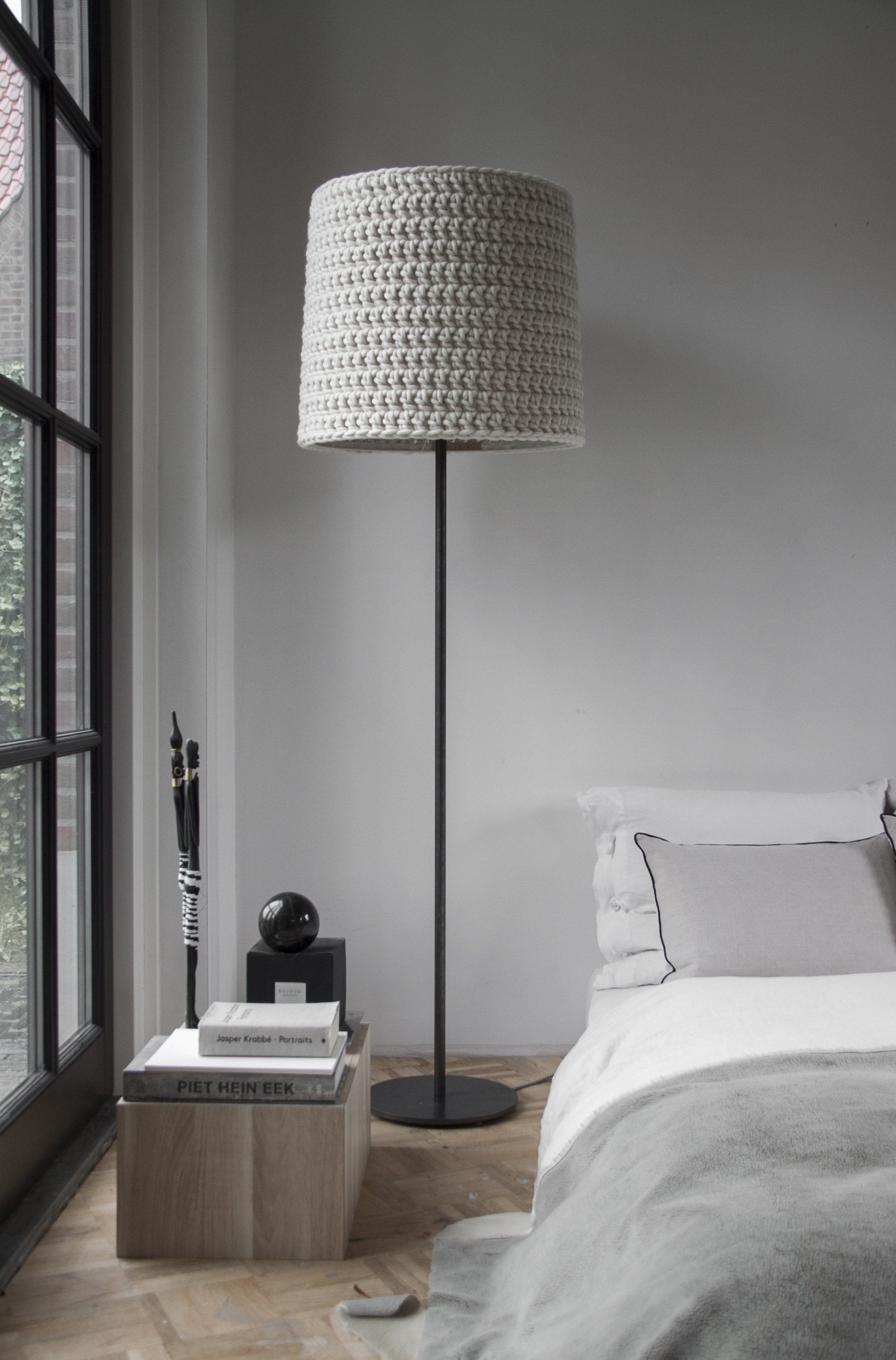 A modern bedroom with a white bed, black floor lamp with a woven lampshade, a wooden side table with stacked books, and large windows with black frames.