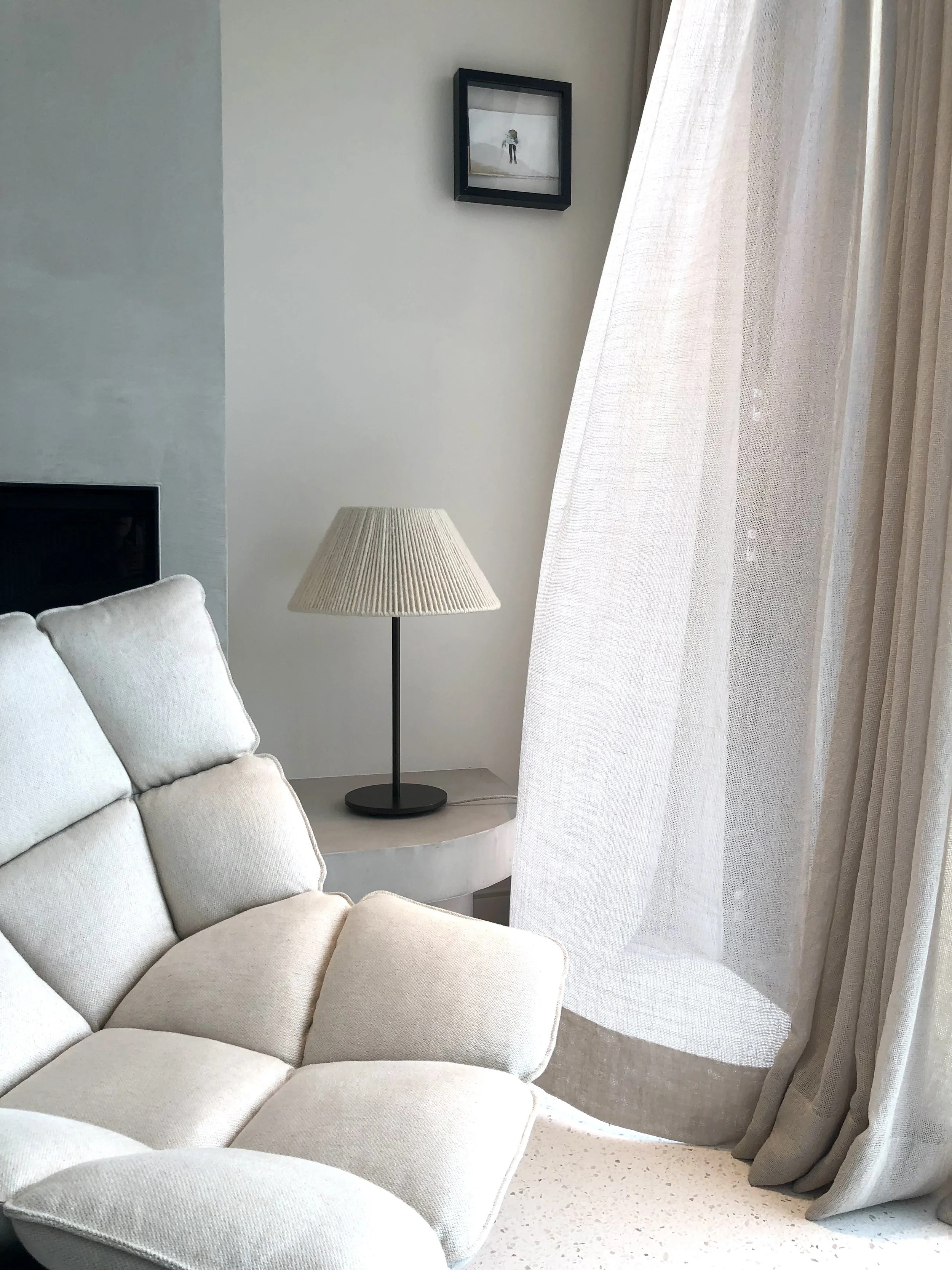 Living room with a white armchair, a side table with a table lamp, white curtains, a wall frame, and a section of a television.
