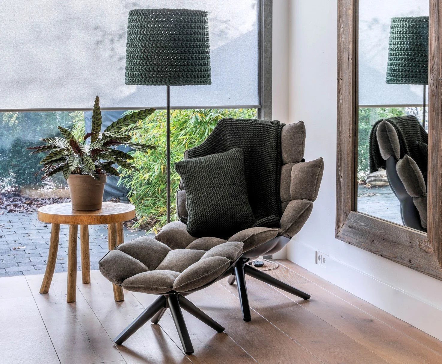A cozy living room corner with a modern recliner chair, a black throw blanket and matching pillow. A round wooden side table with a potted plant. Large windows with blinds and a mirror reflecting part of the chair.