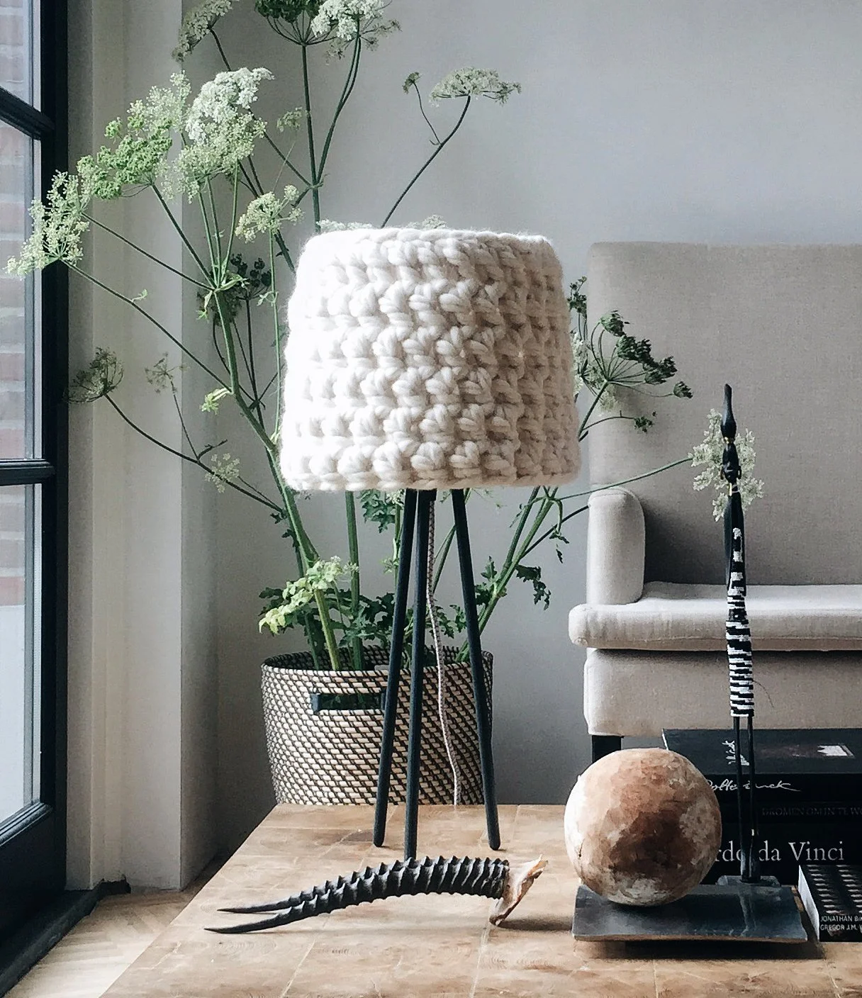 Decorative table setting inside a home featuring a large potted plant with white flowers, a wooden table with a textured white lampshade on a tripod stand, a striped giraffe sculpture, and books including one titled 'da Vinci'