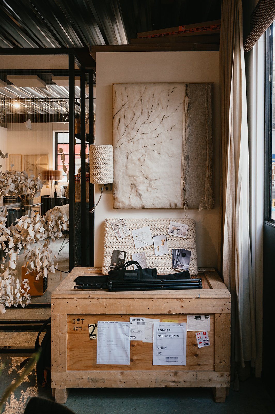 Interior of an art studio or workspace with a large abstract textured canvas on the wall, a wooden crate used as a table with packed shipping labels, some papers, and a black tripod or stand on top. There is a woven wall lamp and various decor and ar