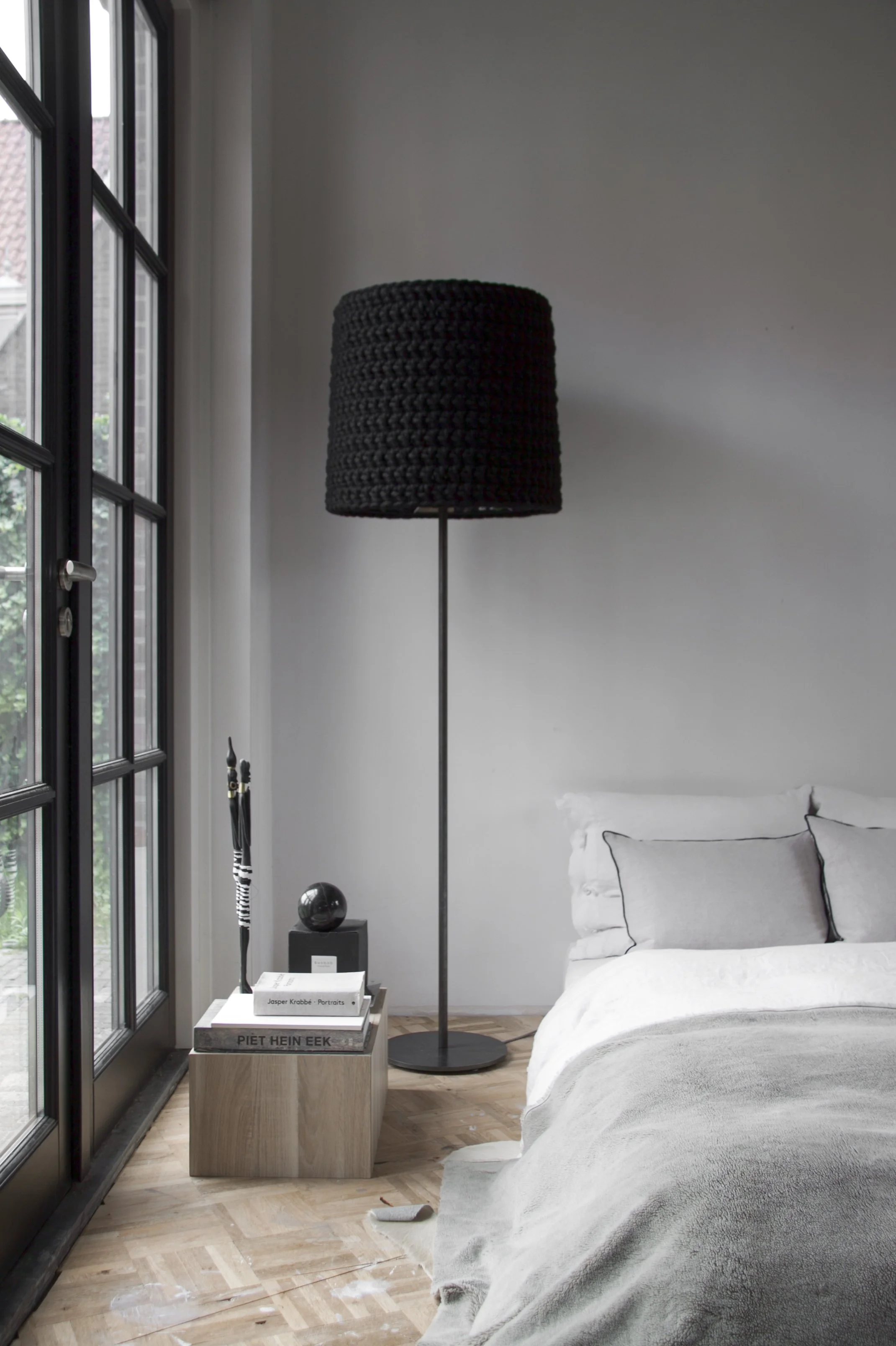 Modern bedroom with a bedside table, books, decorative items, a black woven floor lamp, and an unmade bed with white linens and gray pillows, next to a large window with black frames.