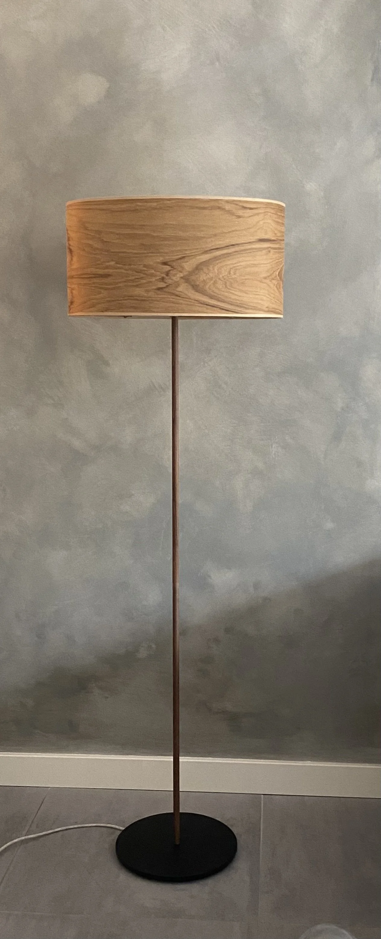 A floor lamp with a wooden lampshade and a round black base, placed against a textured gray wall.