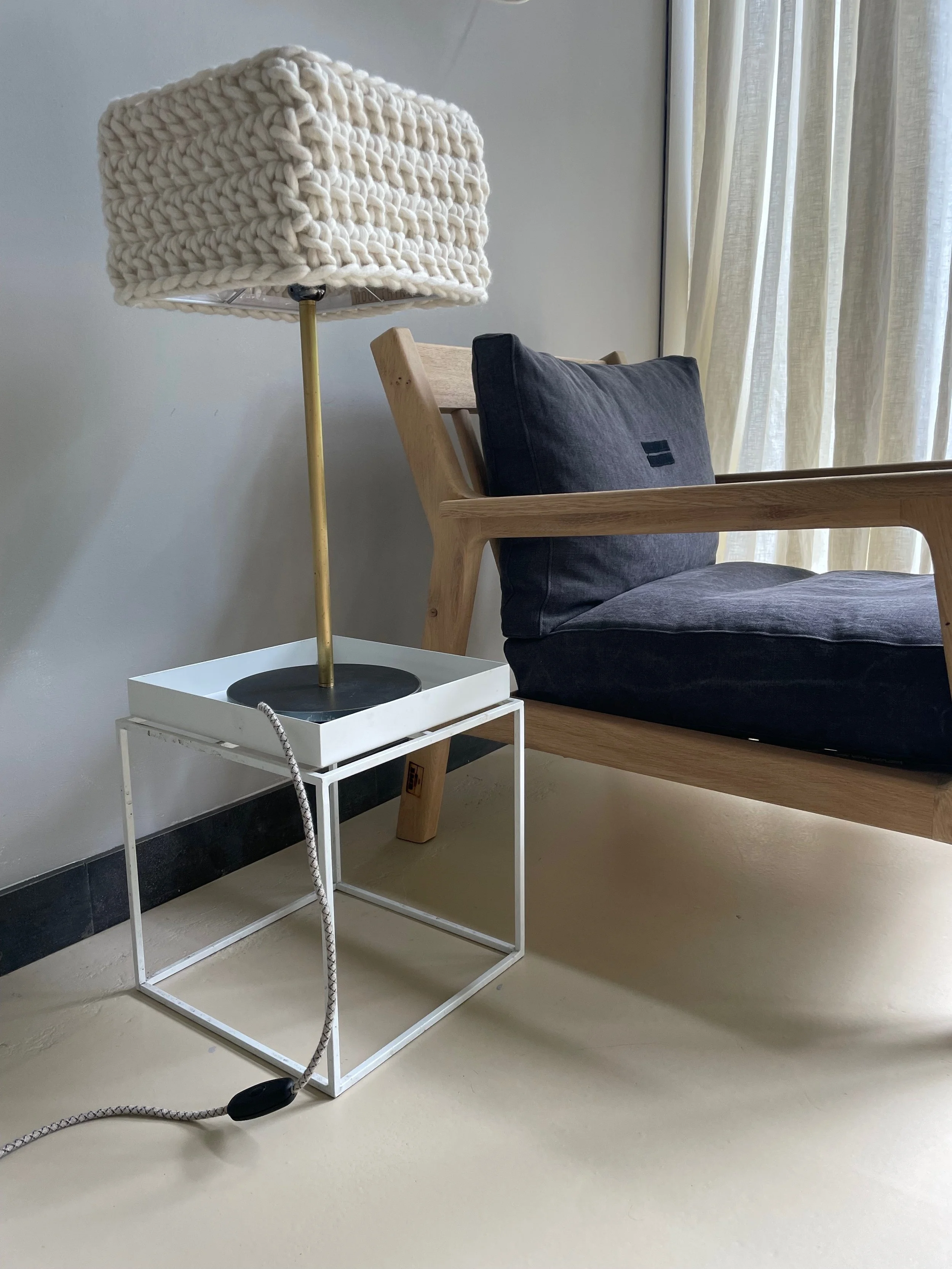 Close-up of a modern nightstand with a lamp on top, sitting next to a wooden chair with a black cushion, near cream-colored curtains.