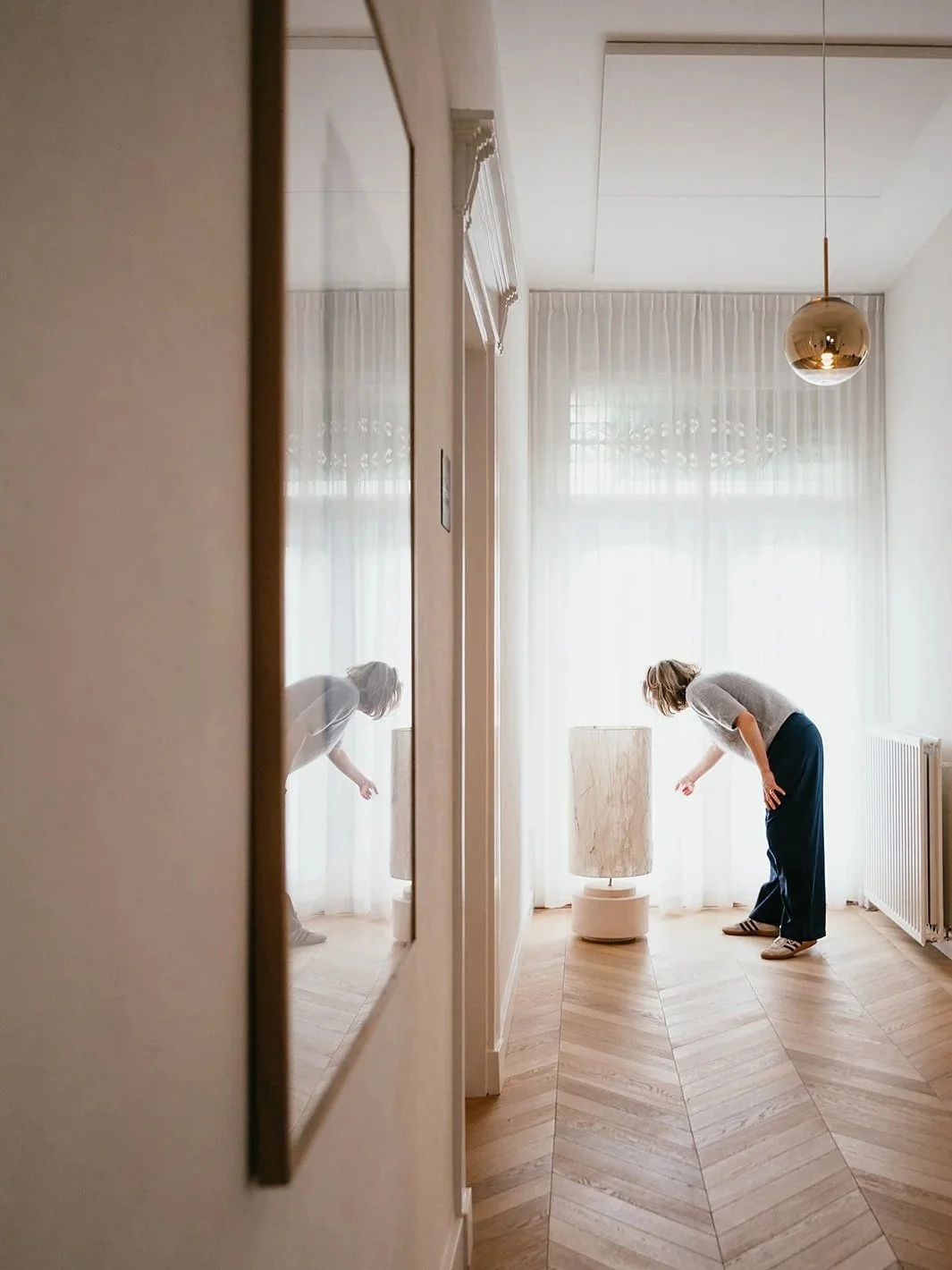 A woman with blonde hair wearing a gray t-shirt, dark pants, and sneakers bent over near a floor lamp in a bright living room with large windows and sheer curtains, with her reflection visible in a mirror on the left side of the image.