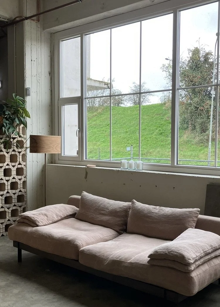 Modern living room with large window showing green grass and trees outside; beige sofa with cushions and folded blankets; wooden floor lamp; decorative wall partition; candles on windowsill.
