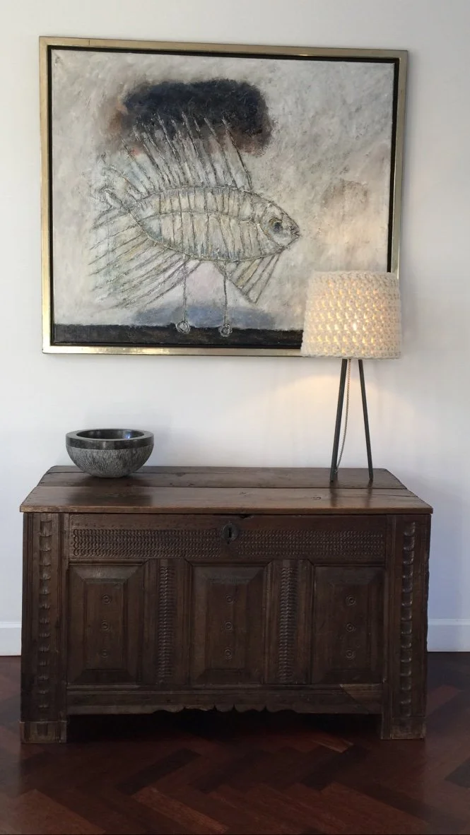 A framed artwork featuring a fish with mechanical or metallic features, hanging above a wooden console table with a ceramic bowl and a standing lamp with a woven shade.