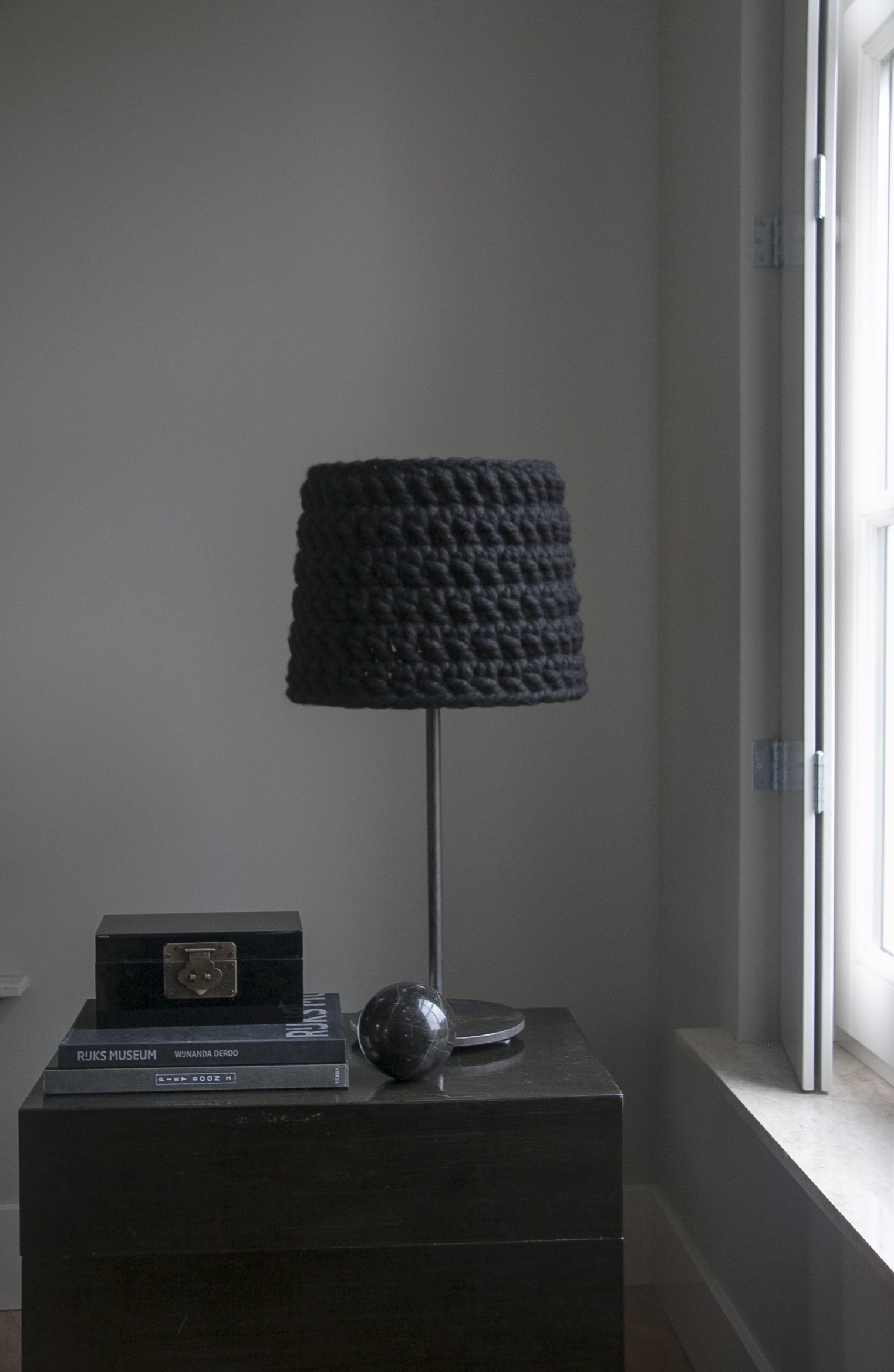 A black woven lampshade on a metal stand sits on top of a black dresser. Next to it are a black box with a latch, a marble sphere, and a round black object. There are a couple of books underneath the box, one titled "RJKS Museum" and another "WJNANDA