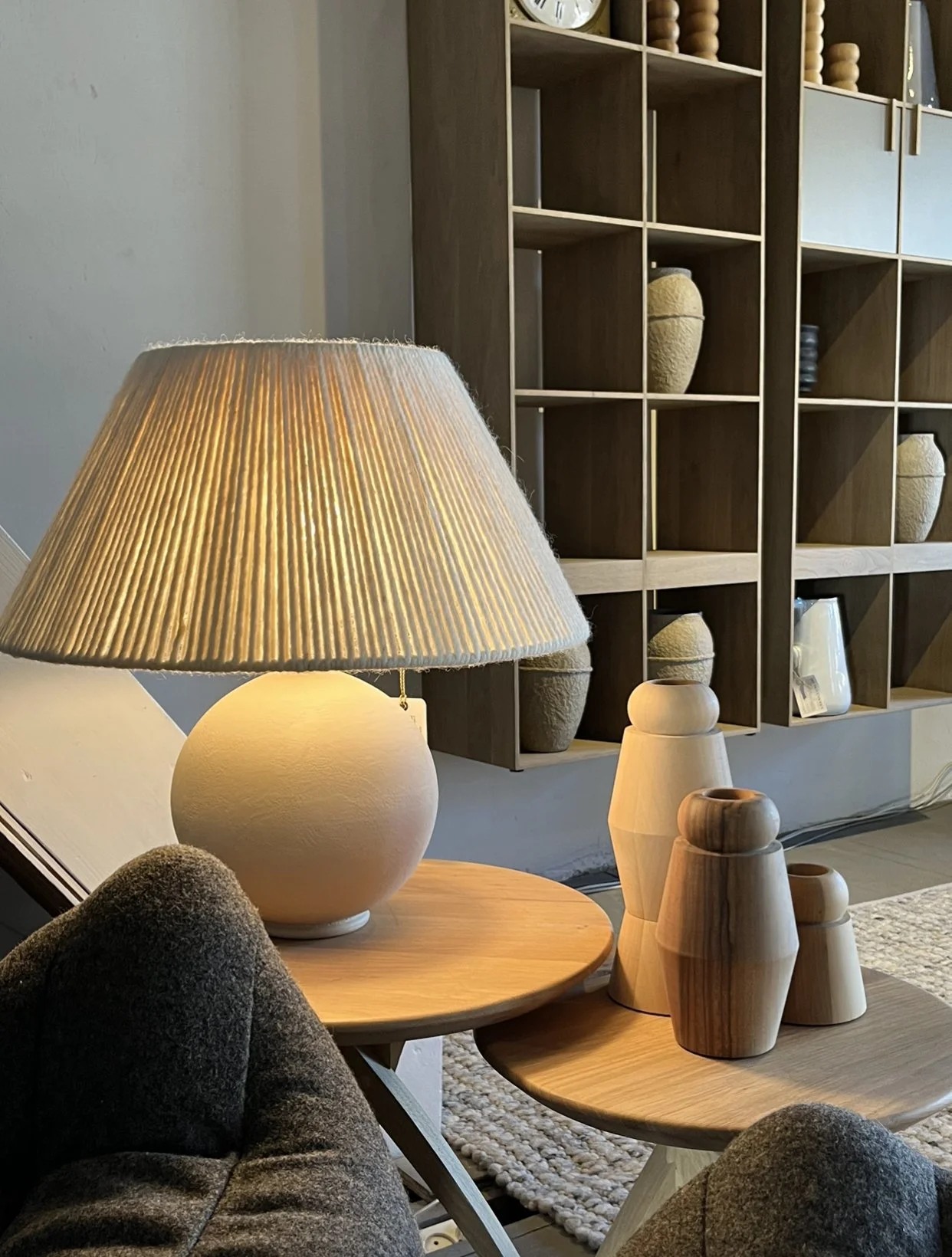 Living room corner with a table lamp, wooden vases, and a neutral-colored sofa. Shelves with vases and decor in the background.