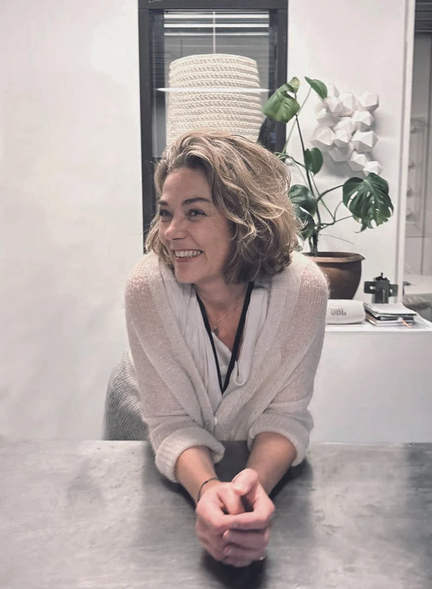 A smiling woman with curly hair seated at a table in an indoor setting with a large potted plant and decorative wall art in the background.