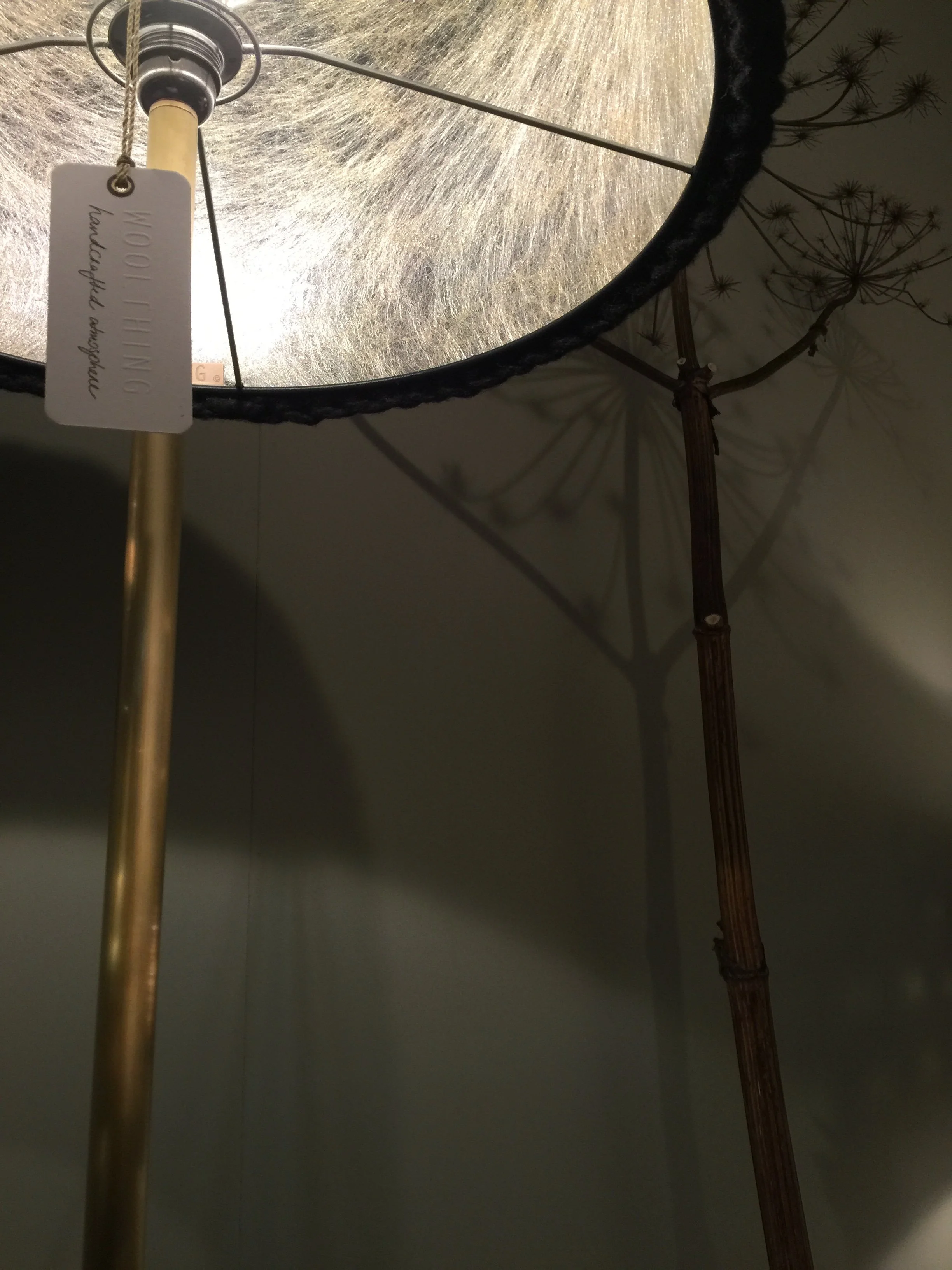 Close-up of a floor lamp with a gold pole, a circular lampshade with a woven pattern, and a tag that reads 'Wood & Linen.' The lamp casts a shadow on the wall behind it.