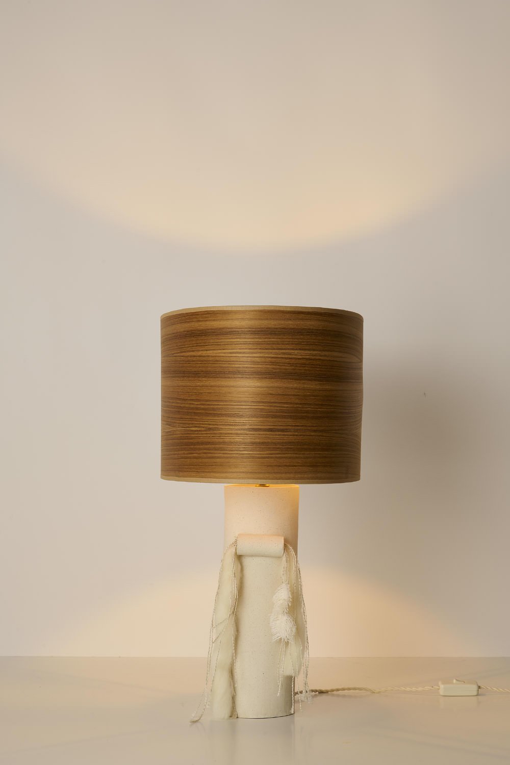 A table lamp with a white ceramic base wrapped in fabric and a wooden cylindrical lampshade emitting light, casting a glow on the wall behind.