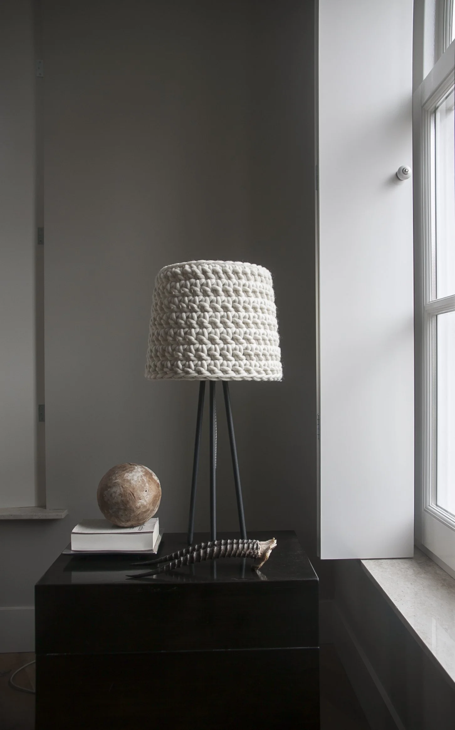 A modern black side table with a macrame lampshade on a tripod stand, a marble sphere, a book, and a decorative shell on top, next to a window with natural light.