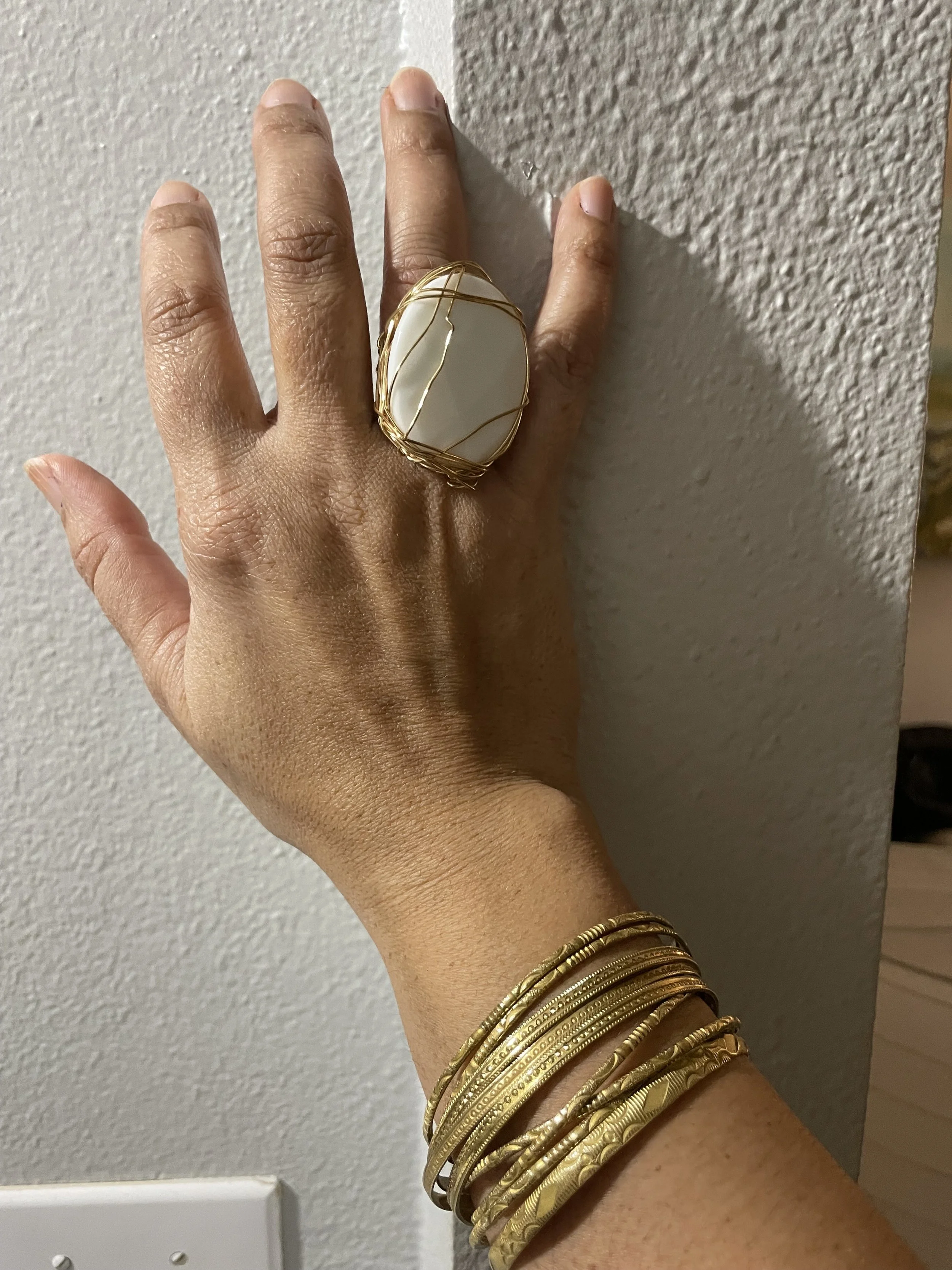 A hand with gold jewelry, including multiple bracelets, a large ring with a white stone wrapped in gold wire, and a plain ring, against a textured wall.