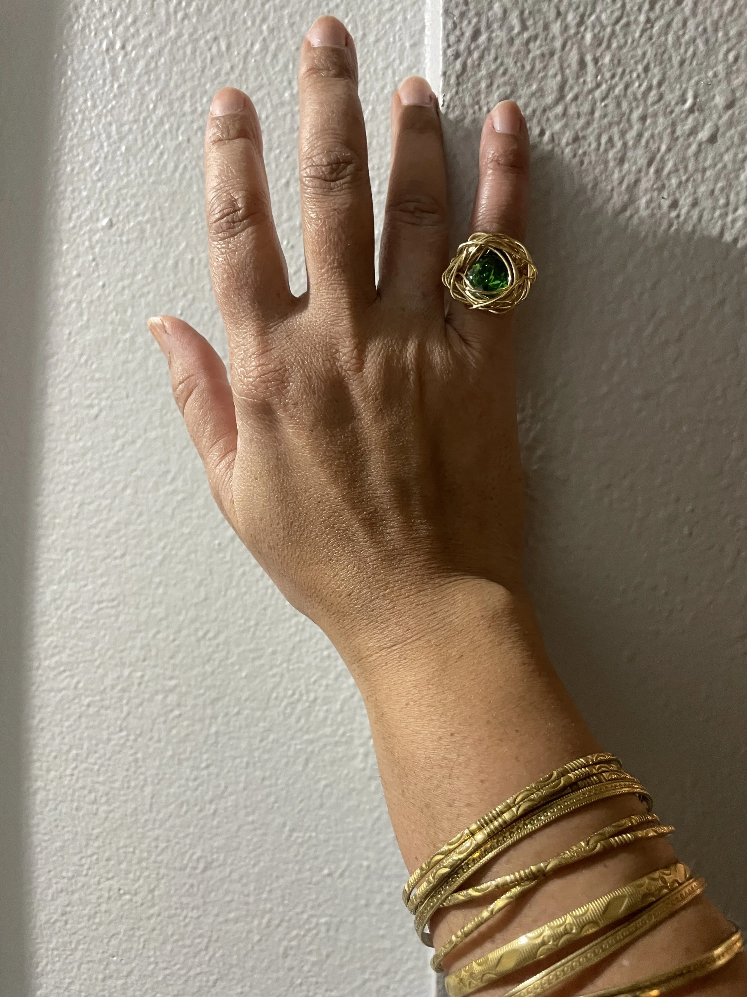 A hand with a large gold ring featuring a green gemstone and several gold bracelets against a textured wall.