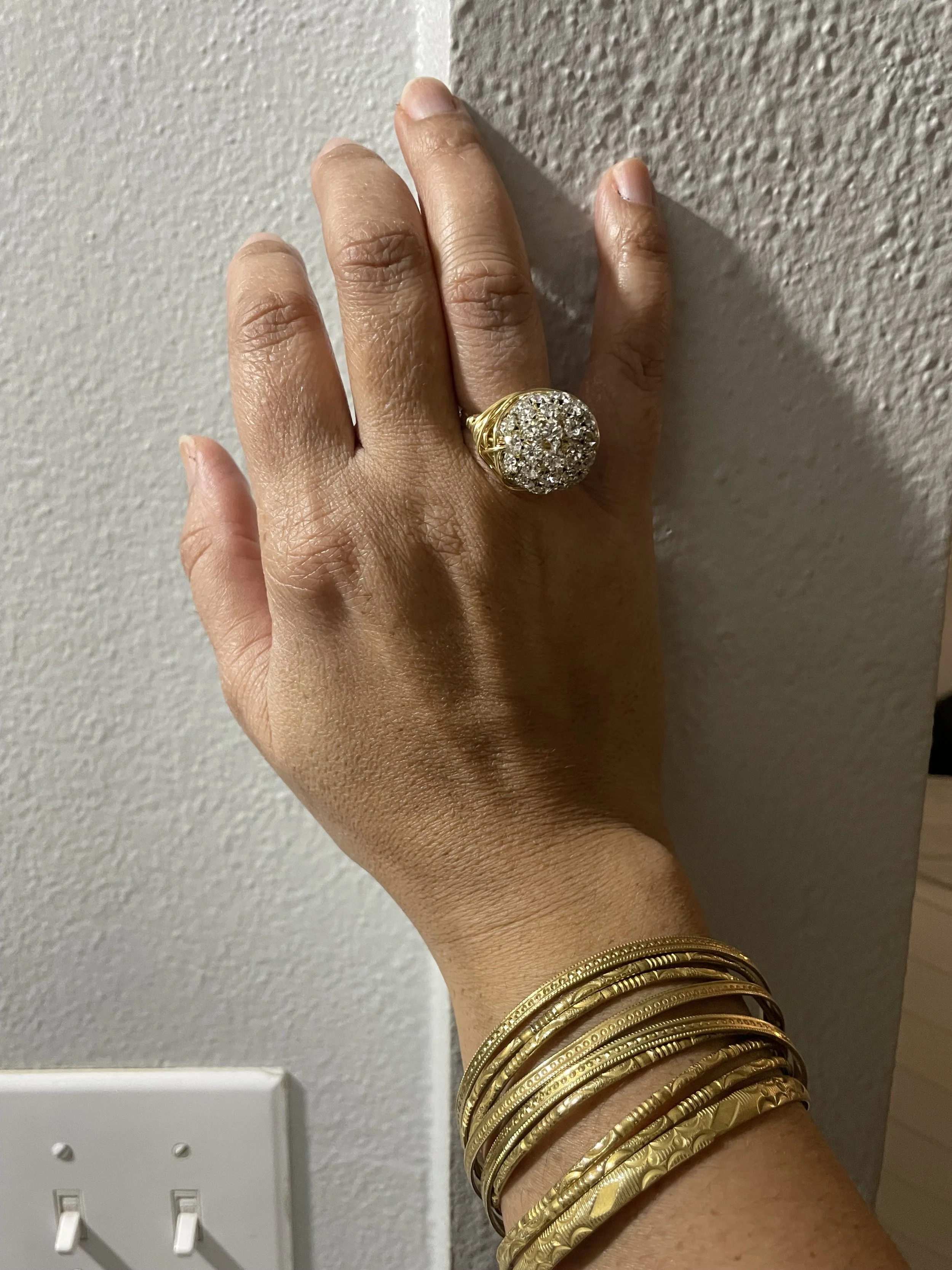 A person's hand is shown resting against a textured wall. The hand wears multiple gold bracelets on the wrist and a large gold ring with a cluster of small diamonds on the middle finger.
