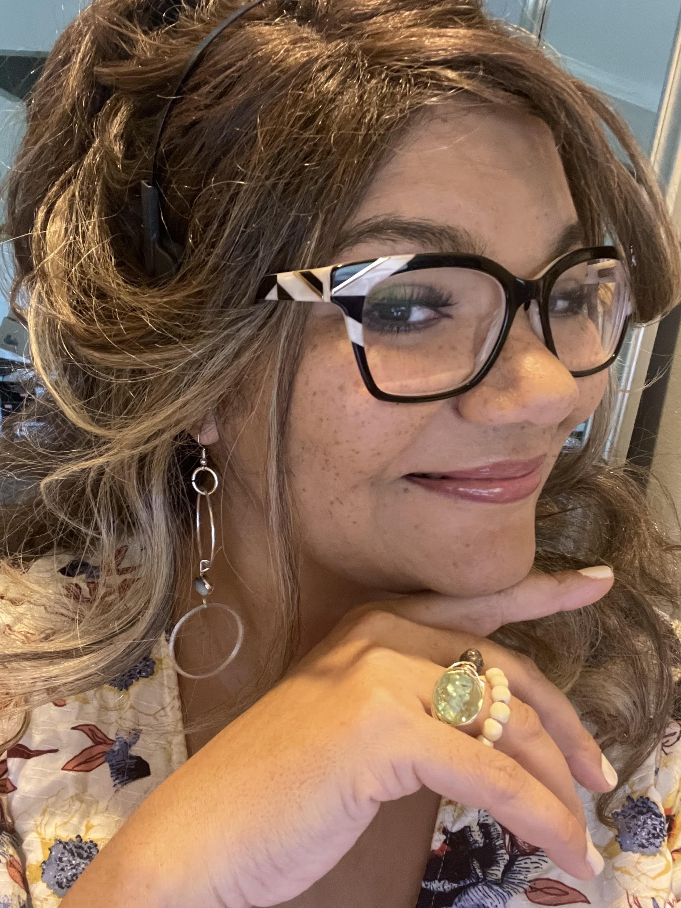 Close-up of a woman with curly light brown hair, wearing glasses, earrings, and rings, smiling and resting her chin on her hand.