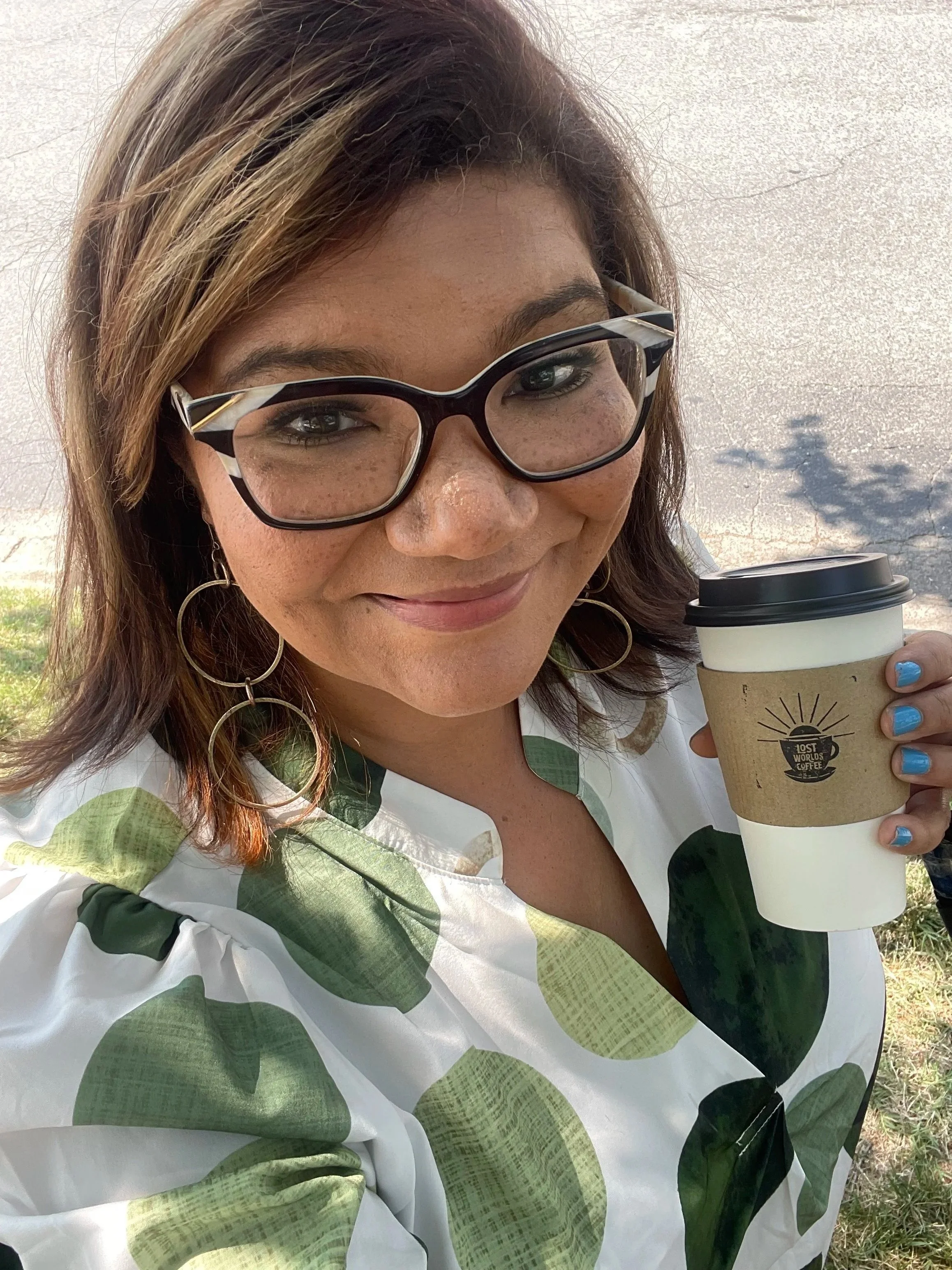A woman with light brown hair, glasses, and hoop earrings, smiling, holding a to-go coffee cup outdoors, wearing a white and green patterned top.