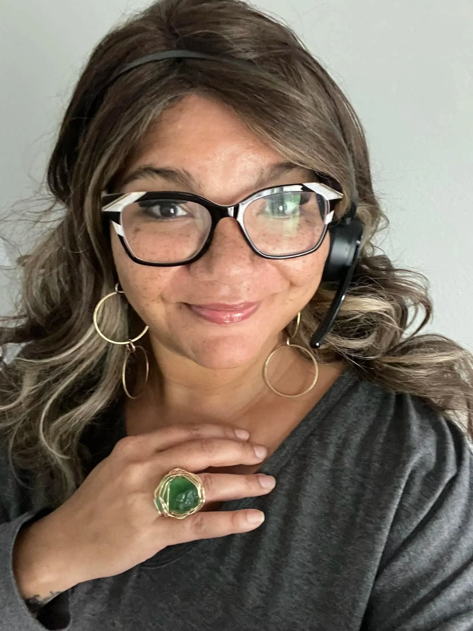 A woman with curly brown hair, glasses, and large hoop earrings smiling, wearing a headset and a gray shirt, with her hand resting on her chest, adorned with a large green ring.