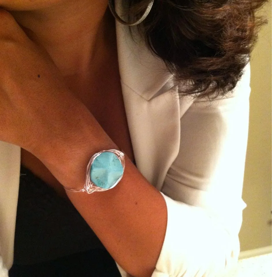 A woman wearing a white blazer and a large silver hoop earring, displaying a bracelet with a large blue gemstone on her wrist.