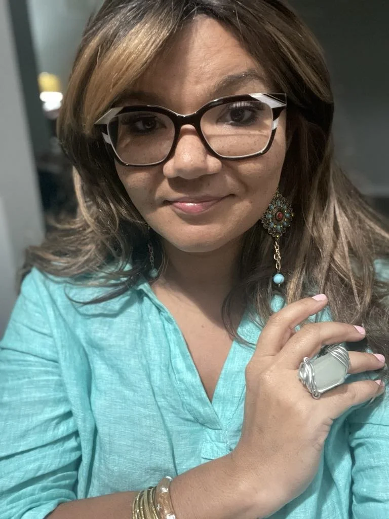 A woman with brown hair, wearing glasses with black and white frames, a turquoise blouse, colorful earrings, and jewelry on her hand and wrist, smiling and looking at the camera.