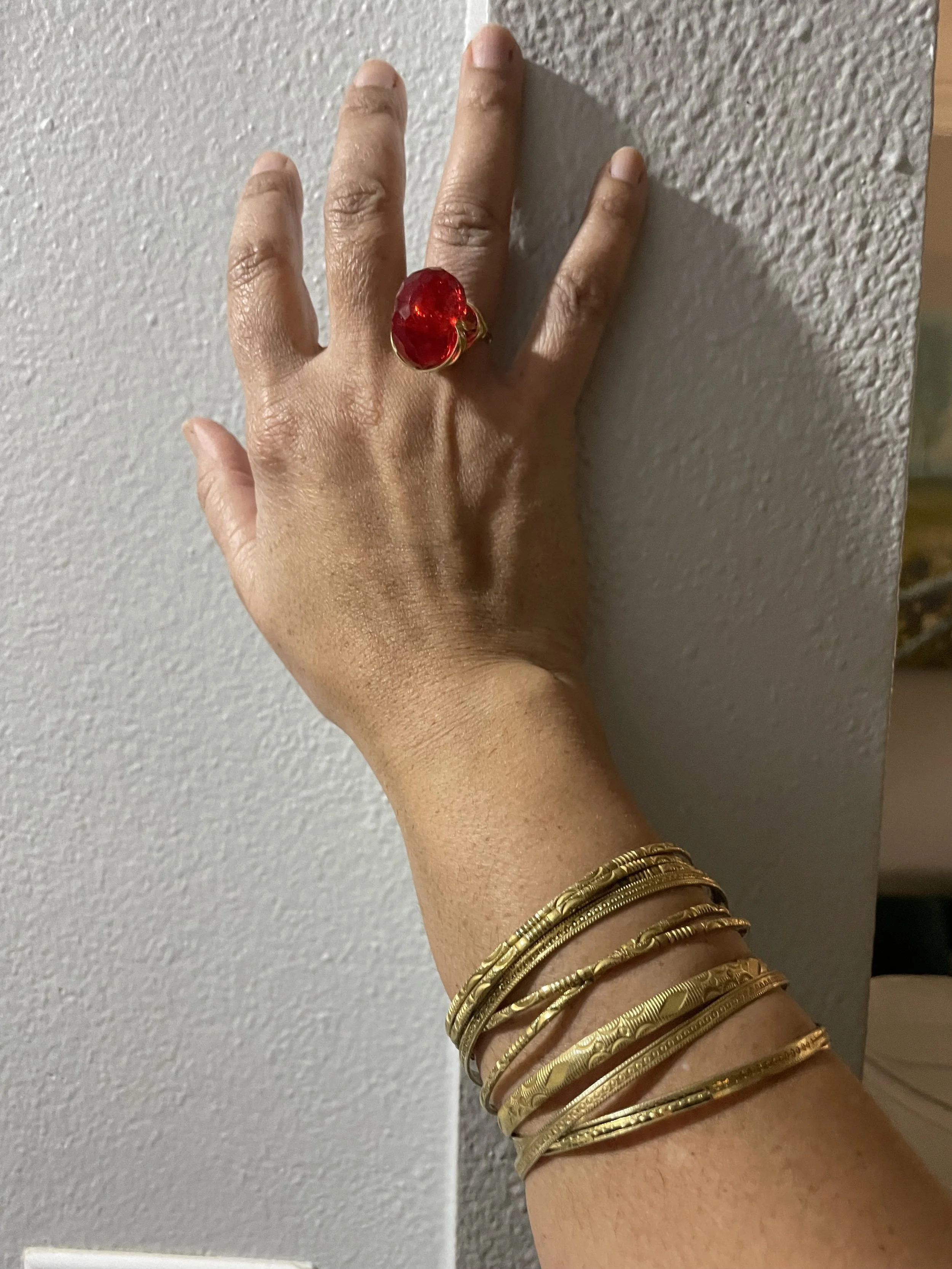 A human hand with multiple gold bracelets on the wrist, a large red ring on the middle finger, and a neutral-colored background.