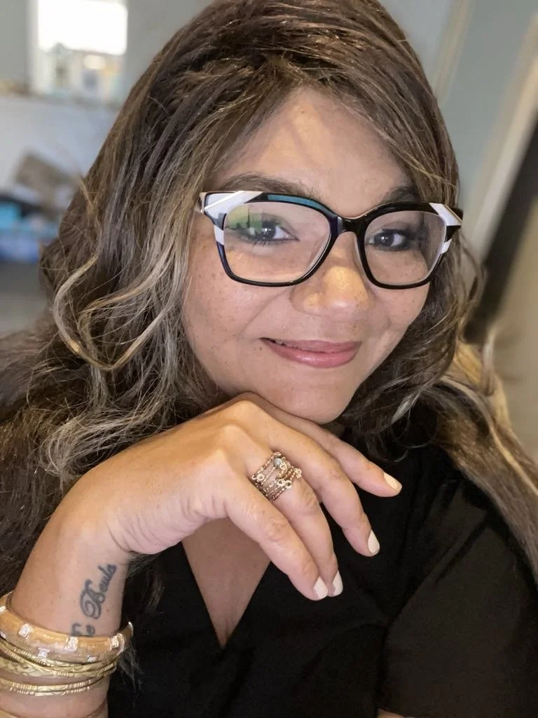 Close-up of a woman with wavy hair, wearing glasses, smiling, resting her chin on her hand, wearing rings and bracelets, and showing a tattoo on her wrist.