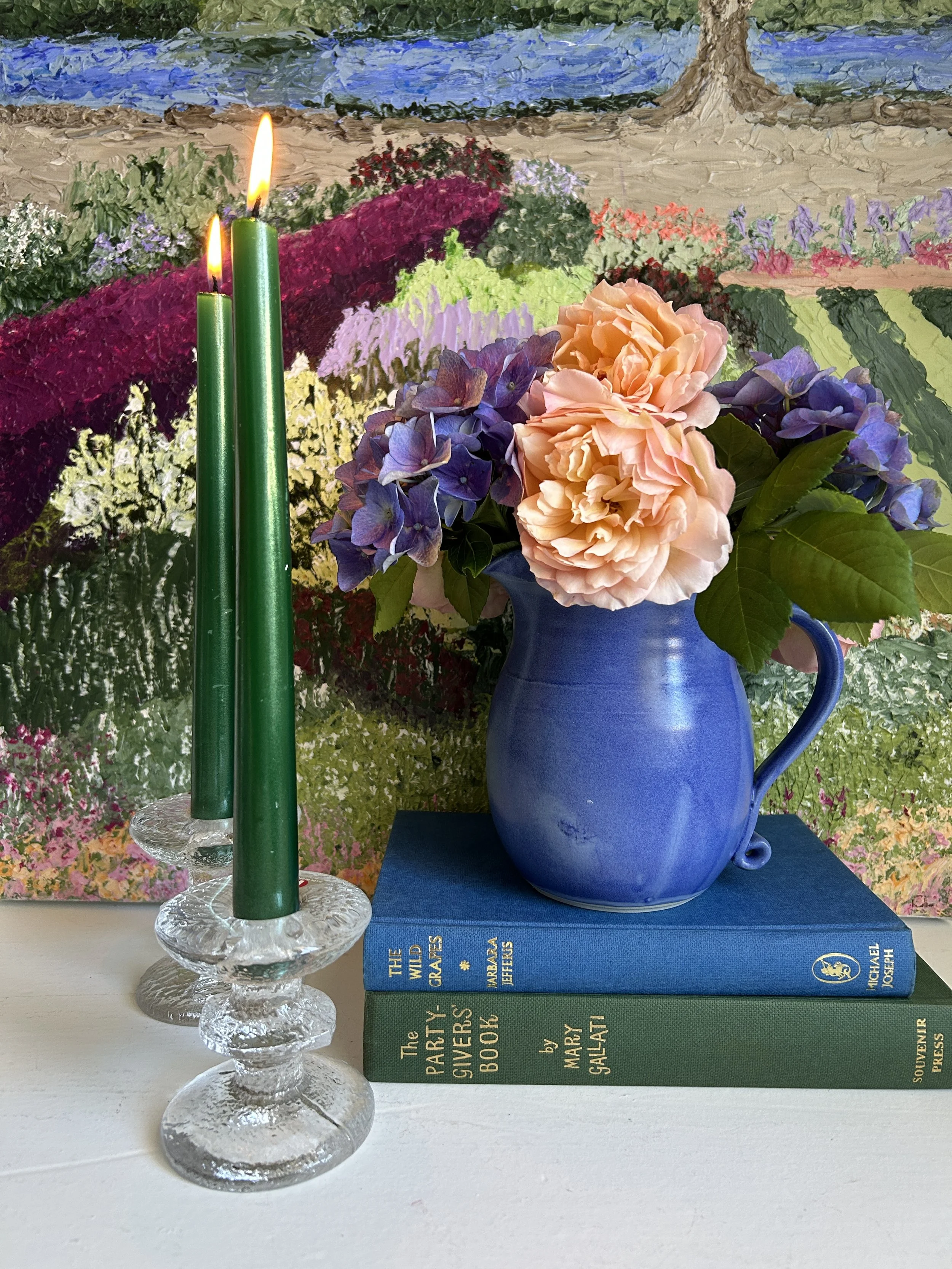 A styled arrangement of two tall green taper candles in textured clear glass candlestick holders beside a blue ceramic jug filled with peach garden roses and purple hydrangeas, resting on stacked vintage books with a colourful garden painting behind.