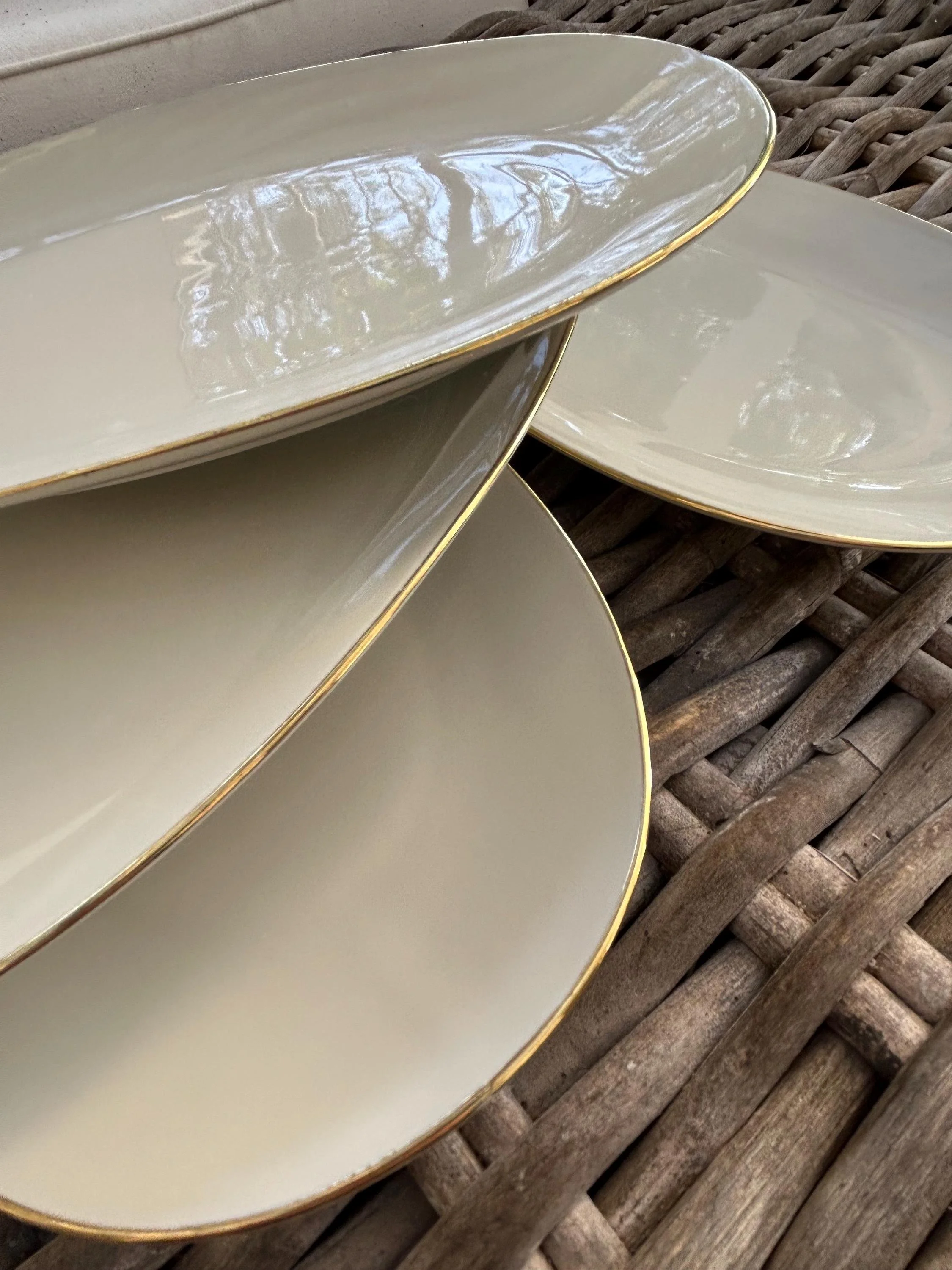 Stack of ivory oval plates with gold trim on woven surface.