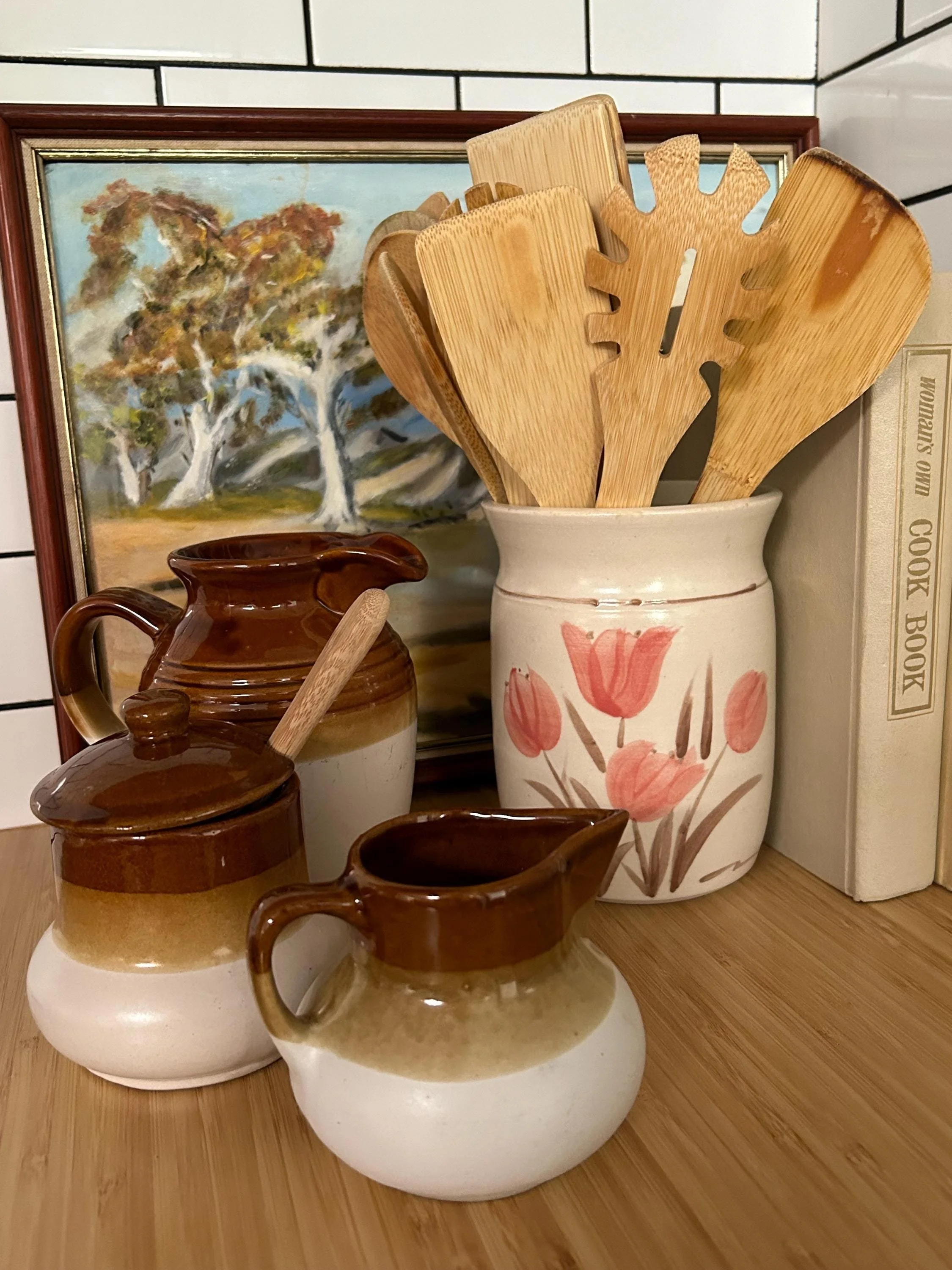 Styled kitchen scene with wooden utensils, books, and vintage stoneware set against tiled backsplash and floral vase